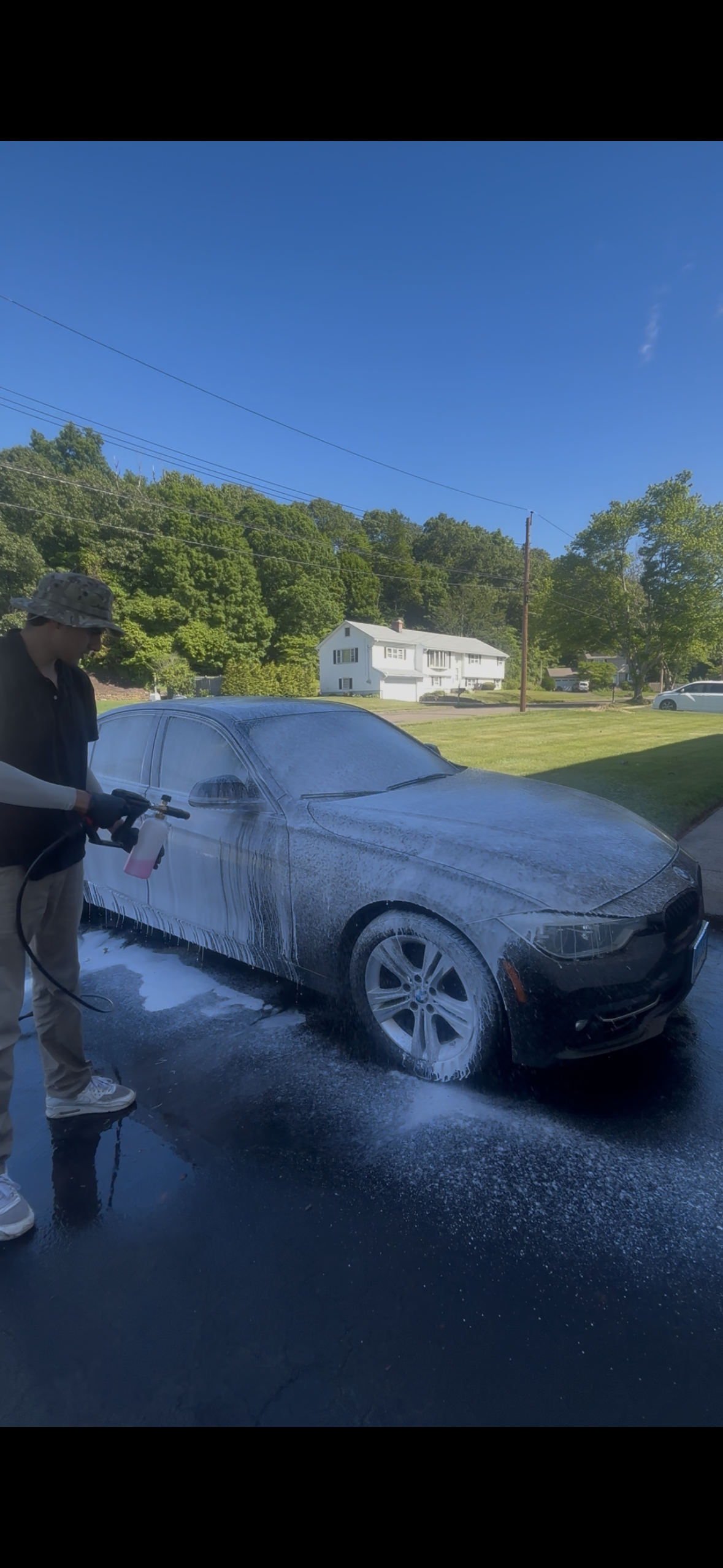 Person using a foam cannon to wash a black car outdoors on a sunny day.