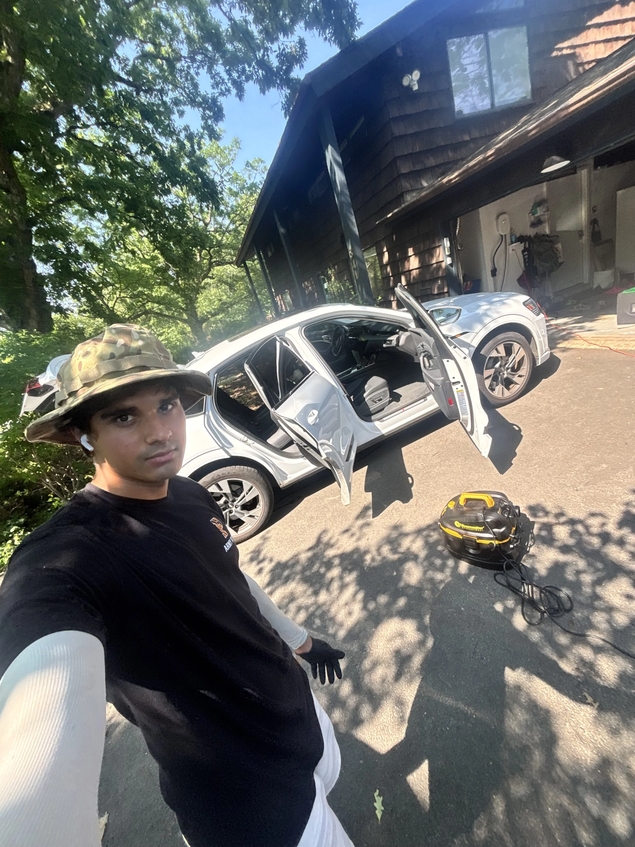 Young man in black and white shirt with a camouflage wide-brim hat taking a selfie outdoors, a partially open white SUV parked on a driveway, tools on the ground, and a black house with a garage and trees in the background.