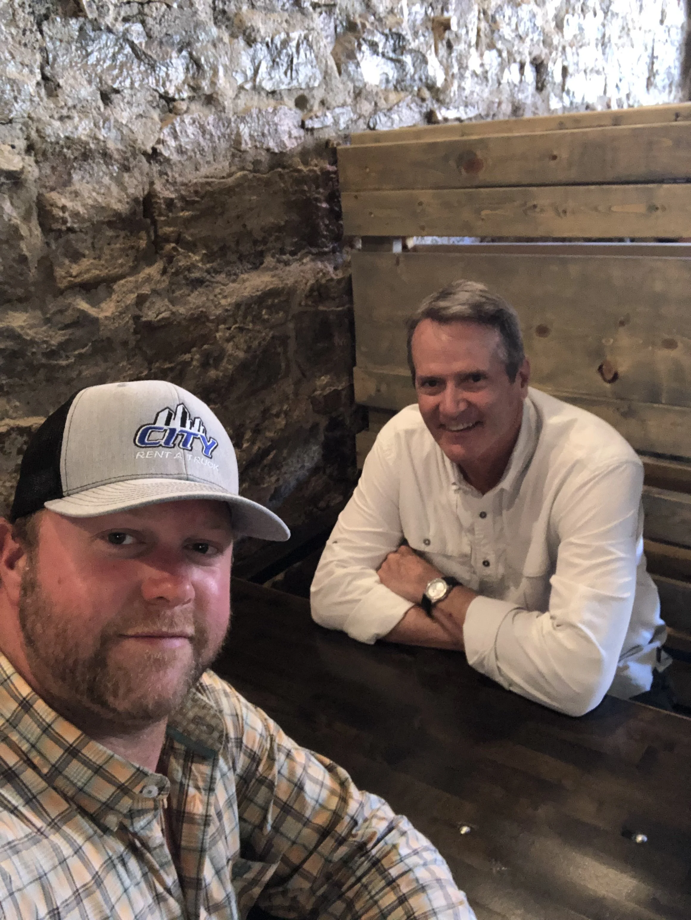 A smiling man wearing a white shirt sitting at a table with a stone wall behind him, and part of another person wearing a baseball cap in front of him.