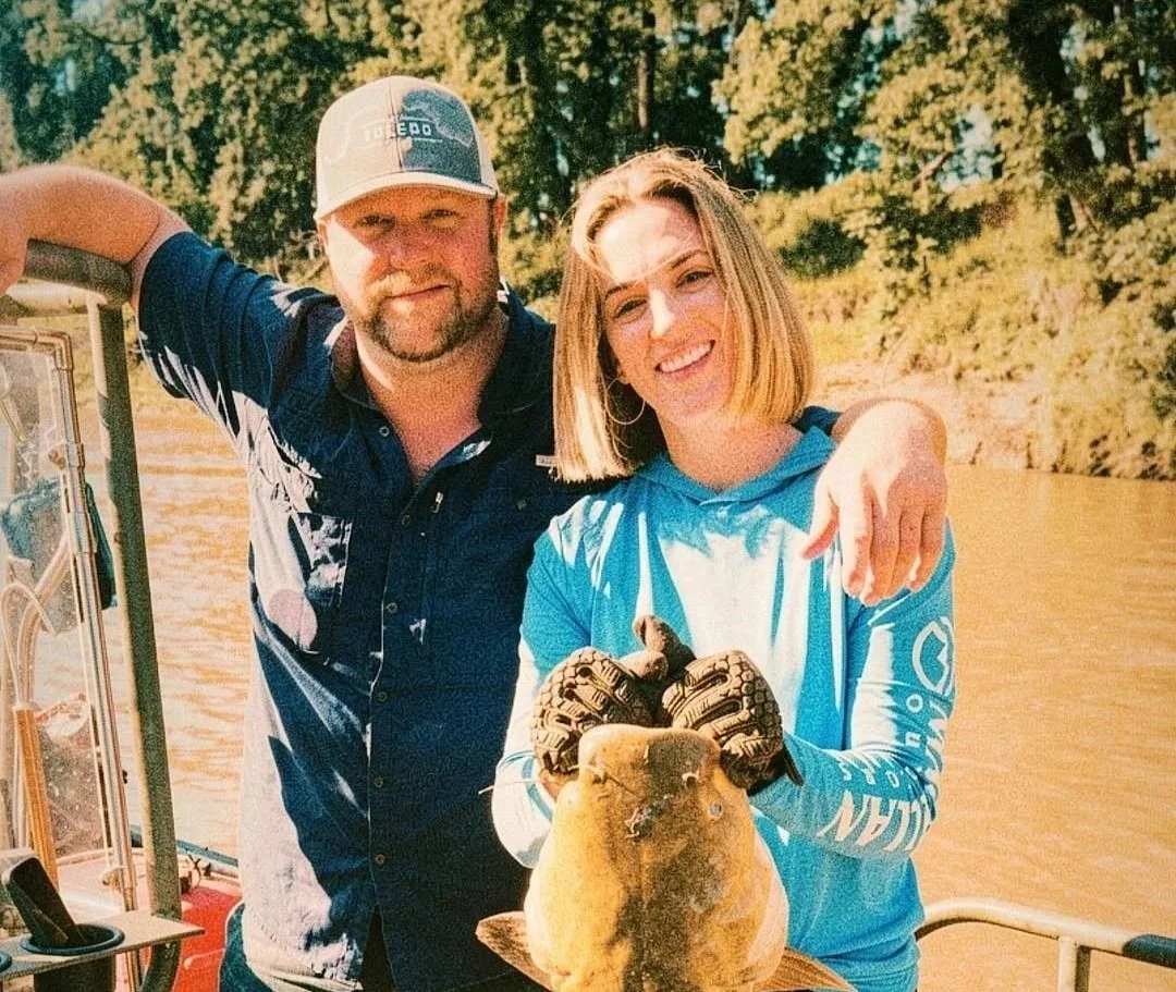 A man and woman on a boat holding a large freshwater fish, with green trees and a river in the background.