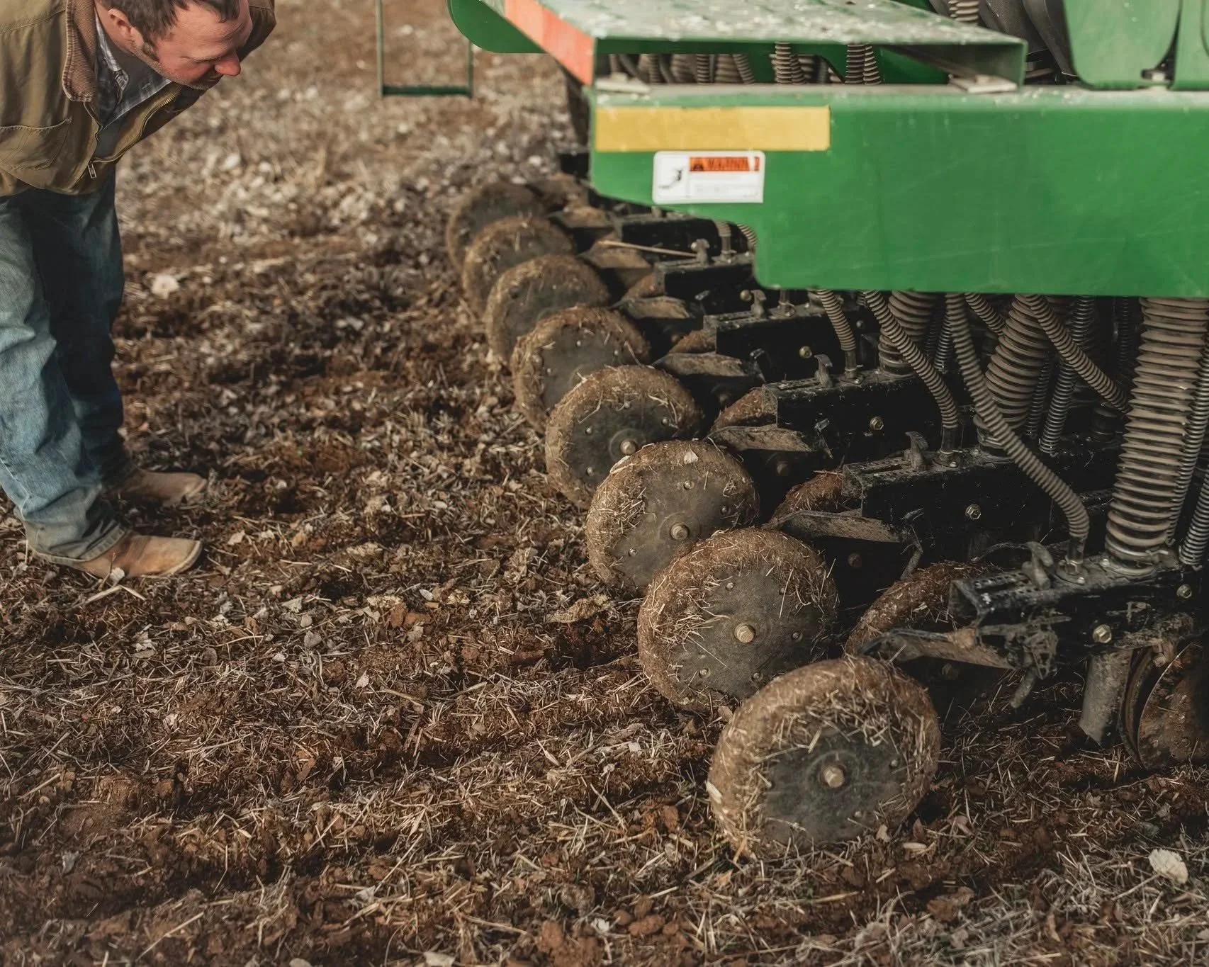 Farmer examining soil with a green tractor attachment in a farm field.
