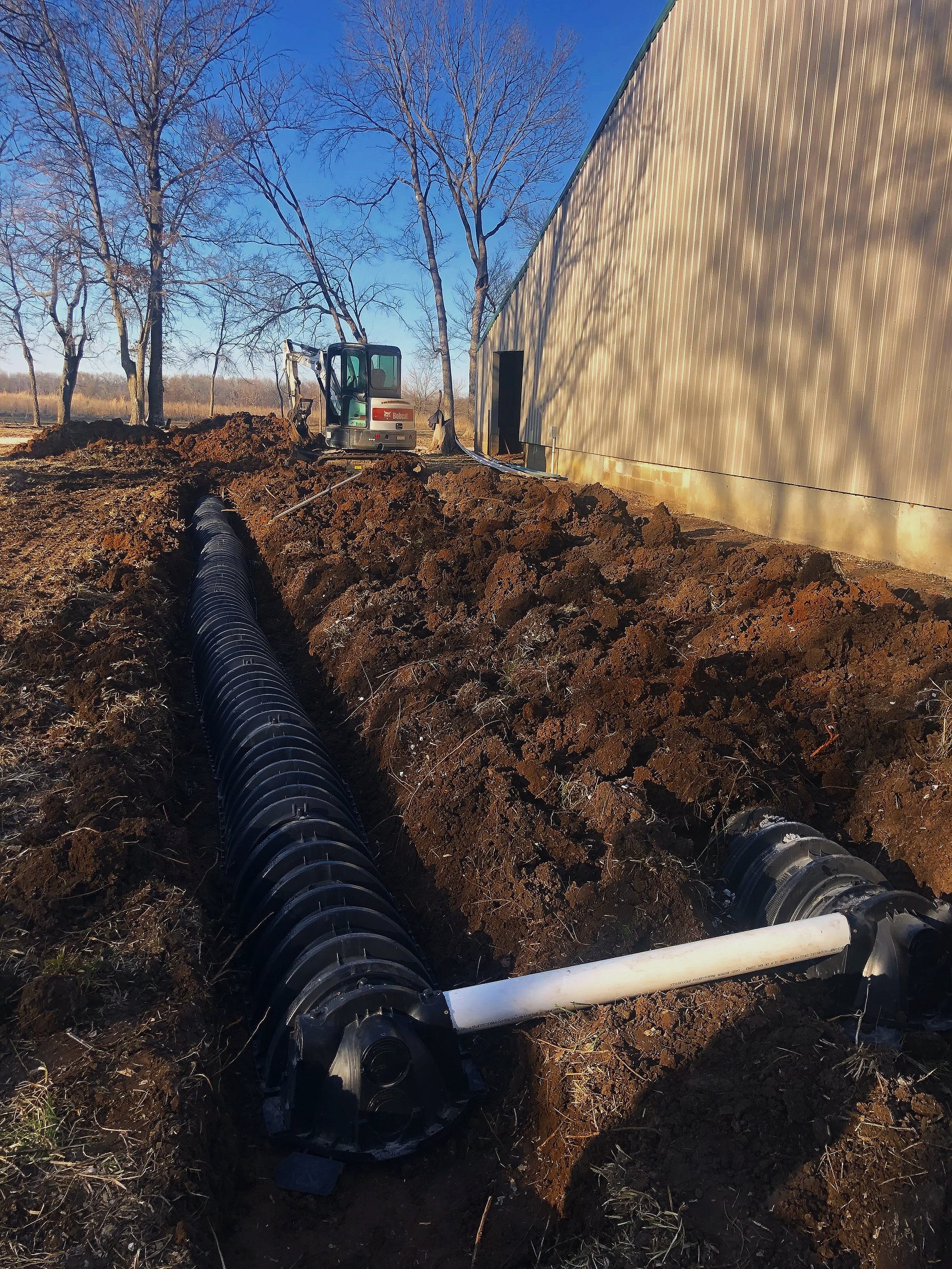 Construction site with a small excavator digging a trench near a large beige metal building, with a large black drainage pipe installed in the trench, and leafless trees in the background under a clear blue sky.