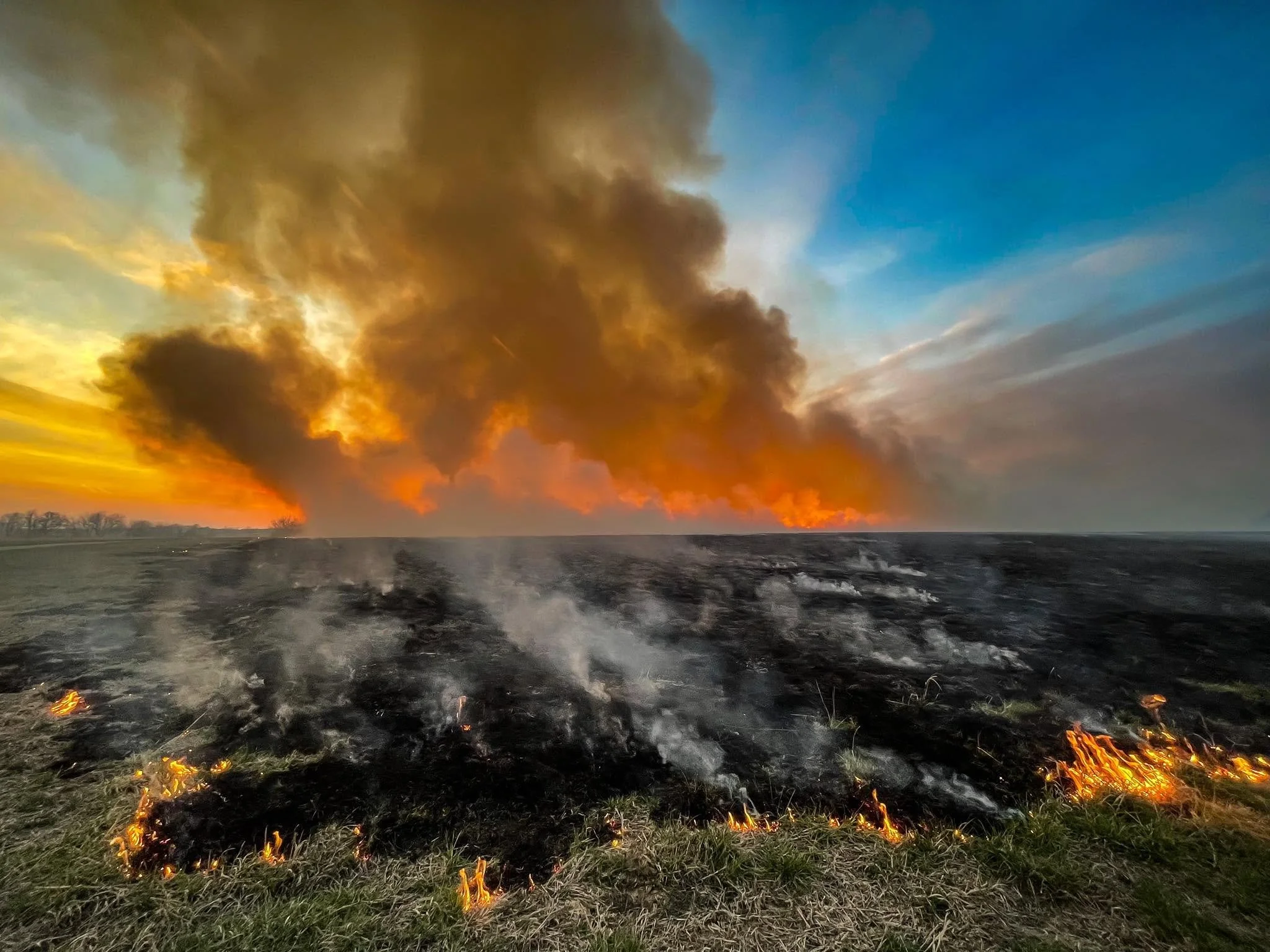Burned grass and soil with small patches of fire in the foreground, smoke rising and a partly cloudy sky during sunset in the background.