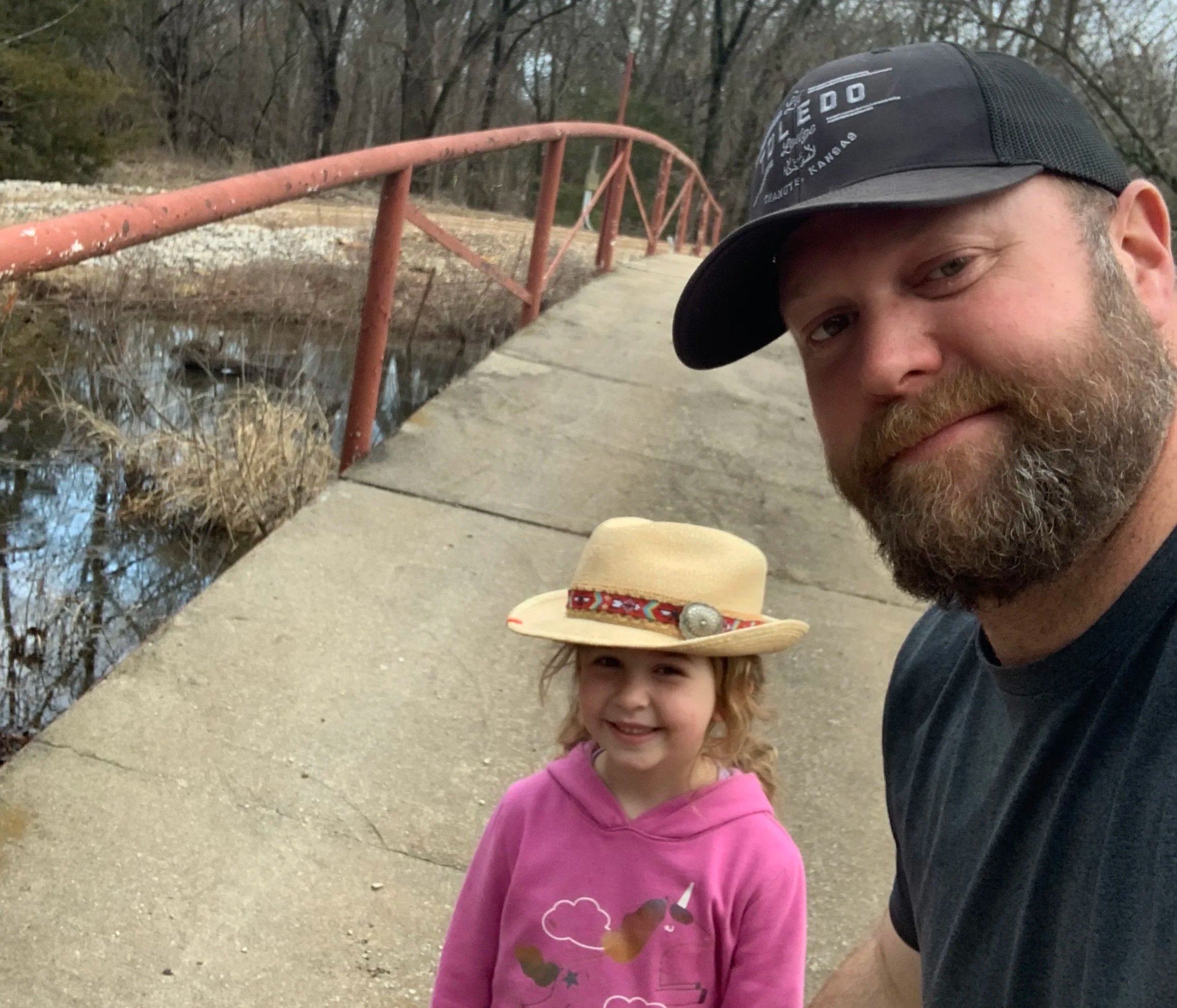 A man and a young girl take a selfie outdoors near a small creek with a rusty red bridge in the background. The man has a beard, wears a black cap, and is smiling. The girl has curly blonde hair, wears a pink hoodie, and is smiling at the camera. The scene appears to be in a wooded area during late afternoon or early evening.