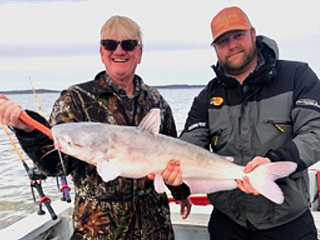 Two men holding a large fish they caught on a boat, with a body of water in the background.