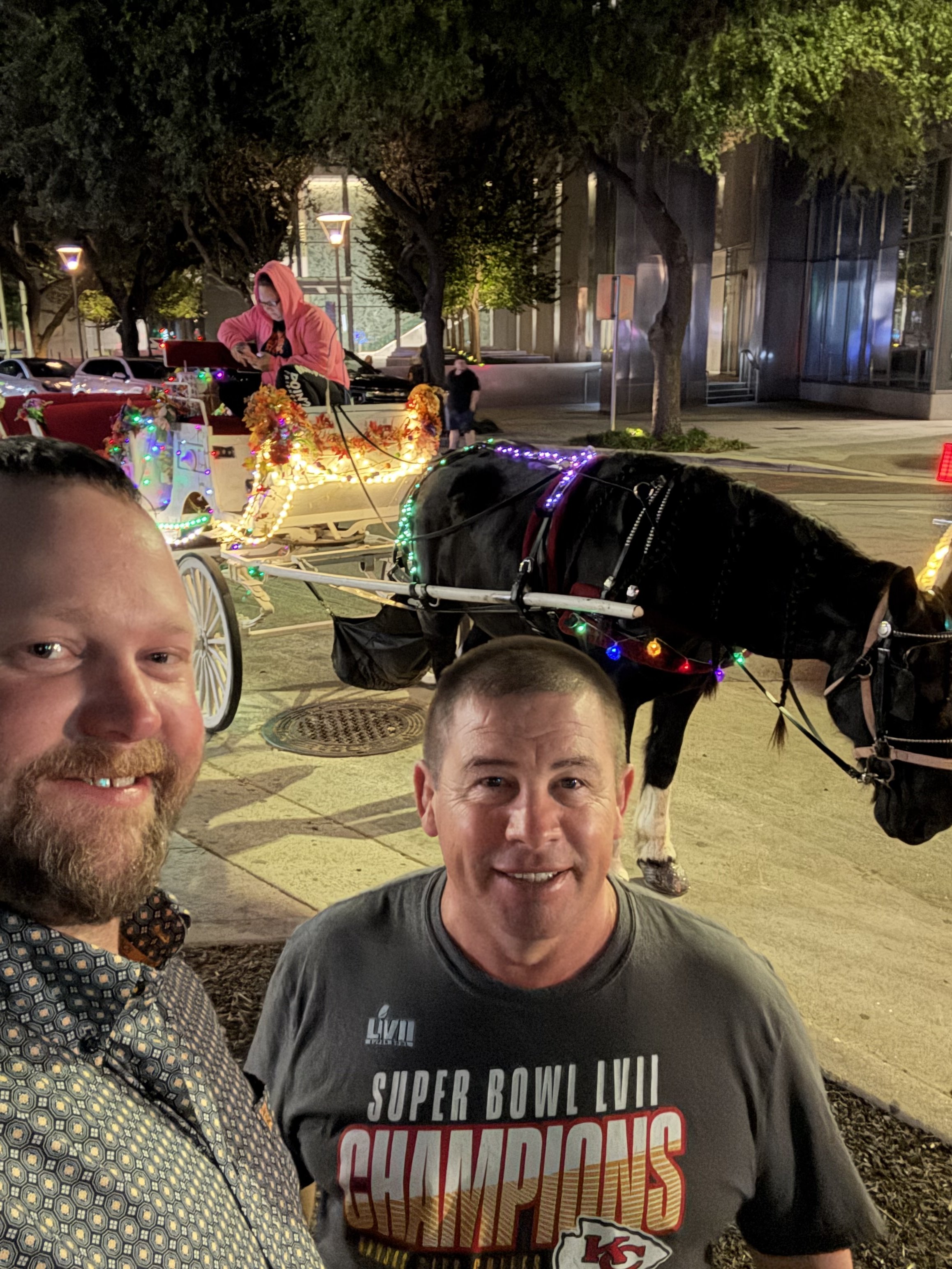 Two men taking a selfie outdoors at night, with a horse-drawn wagon decorated with Christmas lights and a person in a pink hoodie seated inside the wagon visible in the background.