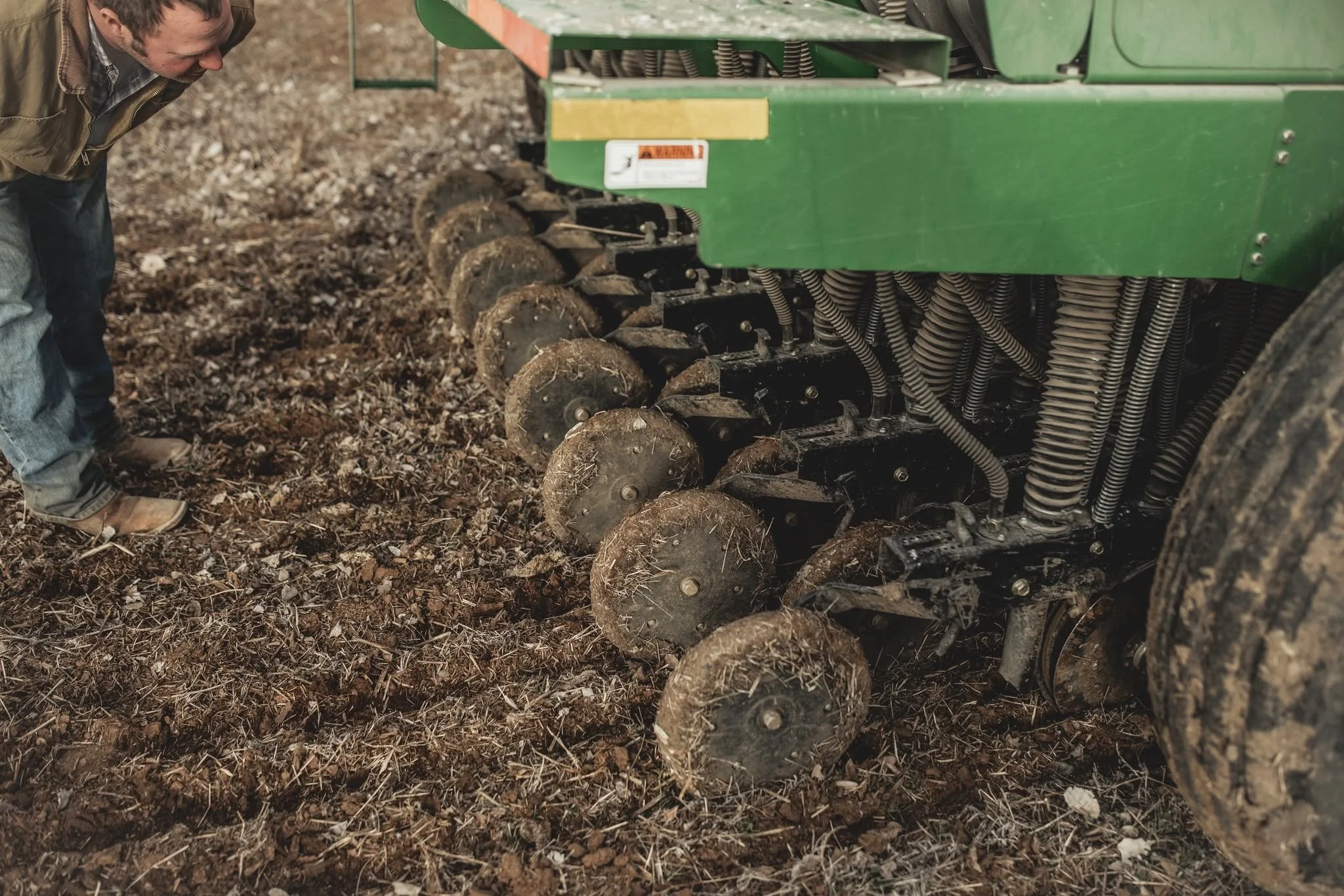 A man wearing a brown jacket and jeans inspecting soil in front of a green vineyard tractor in a field.