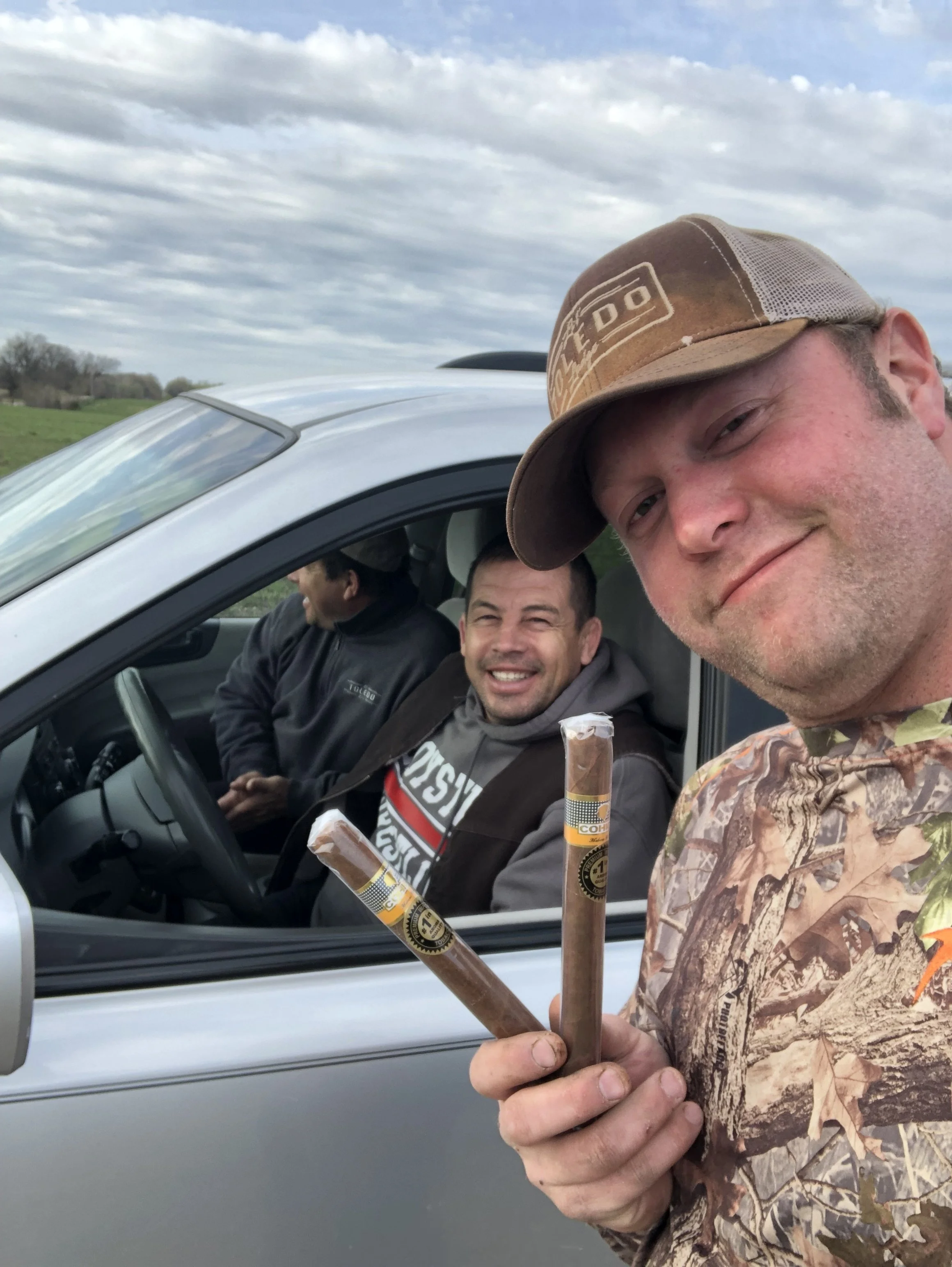 A man wearing a camouflage shirt and a cap holding cigars, standing outside near a silver vehicle, with two other men inside the vehicle, in a rural area with cloudy sky.