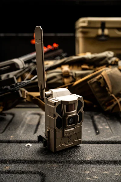 Trail camera with antenna on rugged surface, surrounded by tactical gear and equipment in the background.