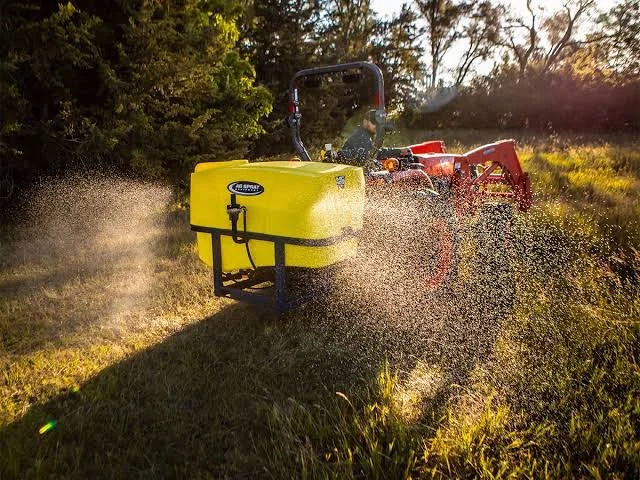 A person operating a tractor with a yellow spray tank attached, spraying water or liquid onto grass in a field during sunset.