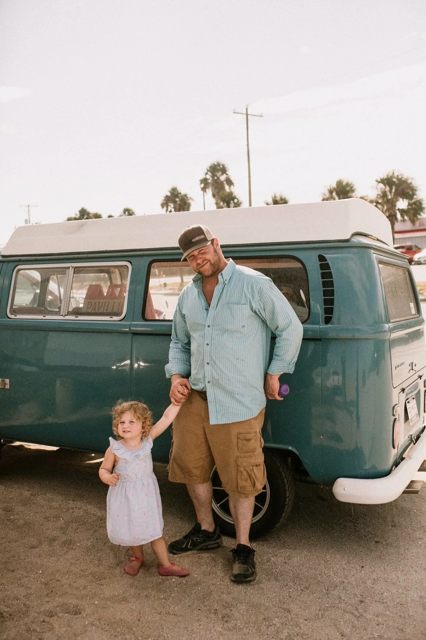 A man with a beard and glasses wearing a baseball cap, light blue striped shirt, and khaki shorts, standing next to a young girl with curly blonde hair in a white dress with pink polka dots, holding her hand. They are standing in front of a teal camper van, with palm trees and utility poles visible in the background on a sunny day.