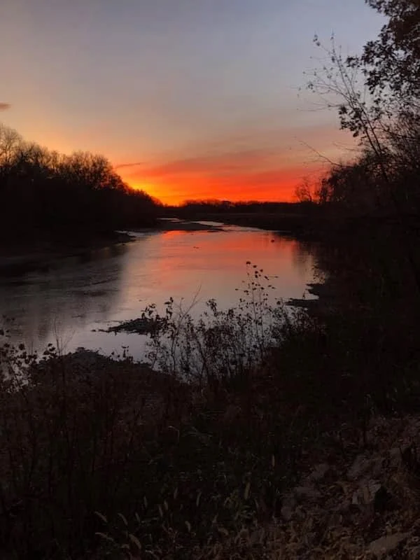 Sunset over a calm river with trees on both sides and colorful sky reflections in the water.