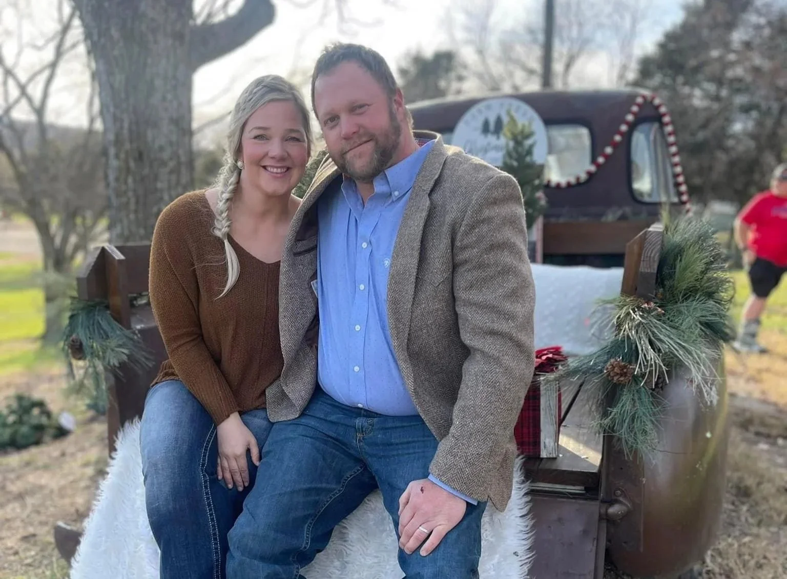 A smiling couple sitting on a decorated wooden bench outdoors. The woman has long blonde hair in a braid, wearing a brown sweater, and the man has short, light brown hair, wearing a blue shirt and a beige jacket. In the background, there is a vintage truck decorated with greenery and holiday ornaments, and a person in a red shirt is walking by.