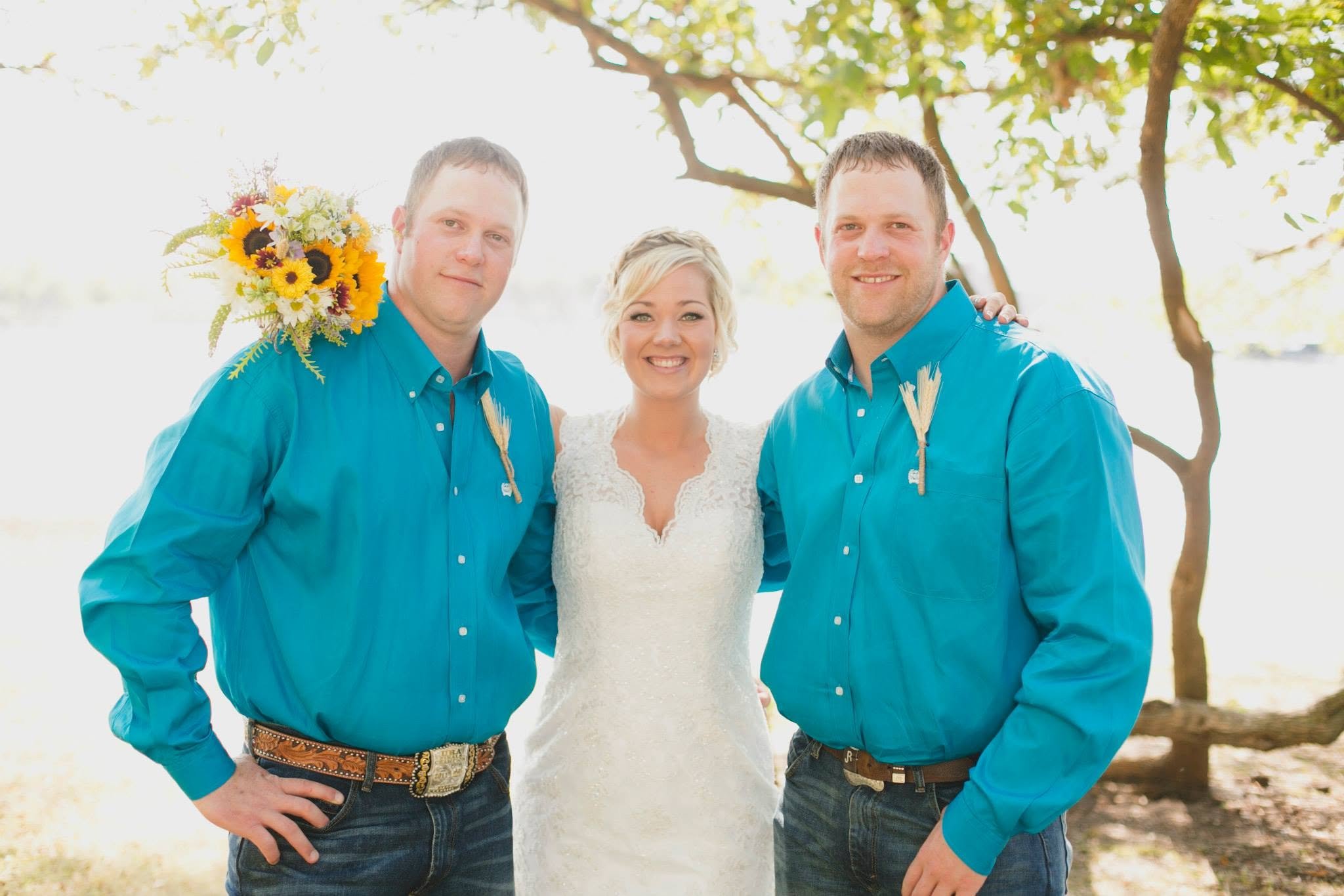 Three people standing outdoors in front of a tree with green leaves, smiling at the camera. The woman is in the middle, wearing a white dress, and two men are on either side, wearing blue shirts. The man on the left is holding a bouquet of sunflowers and other flowers over his shoulder.