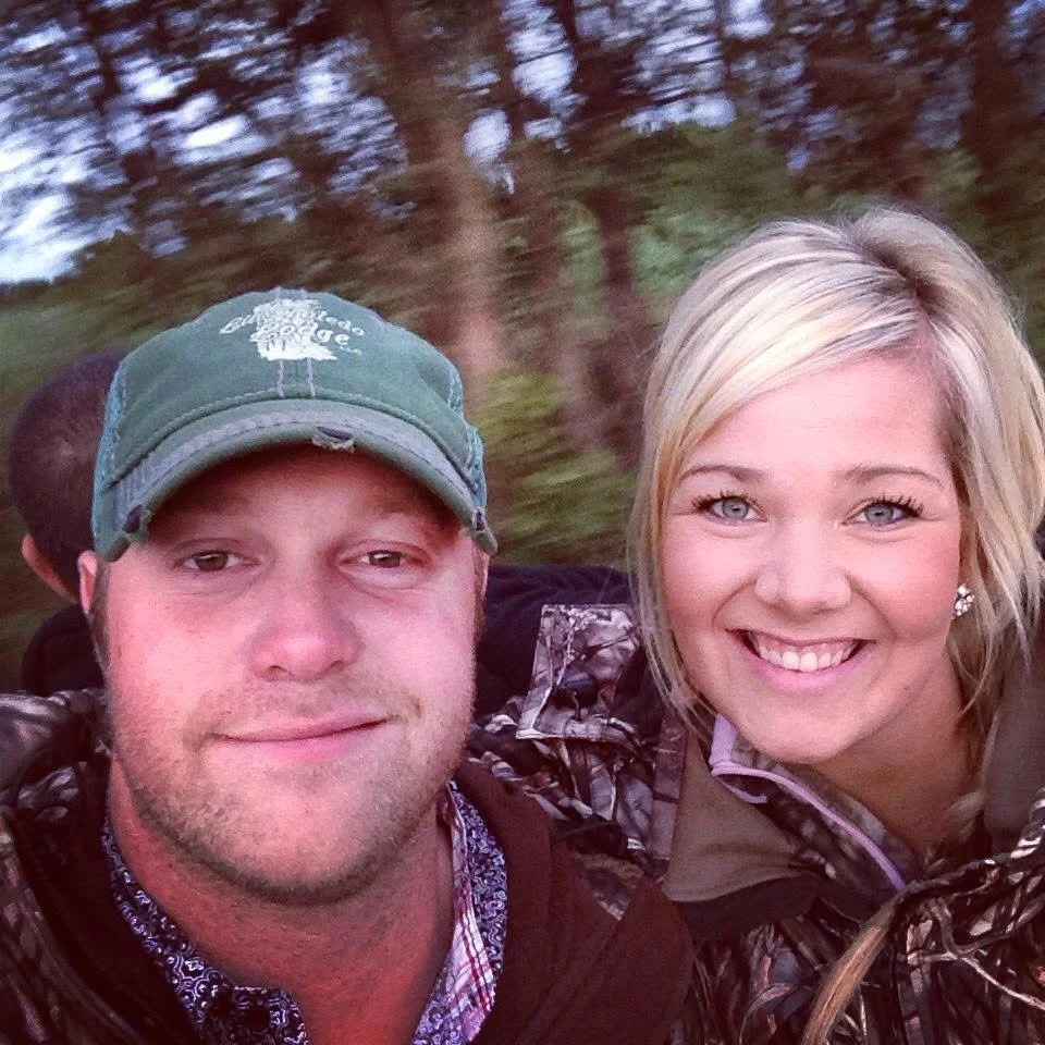 A man and woman taking a selfie outdoors, with trees blurred in the background.