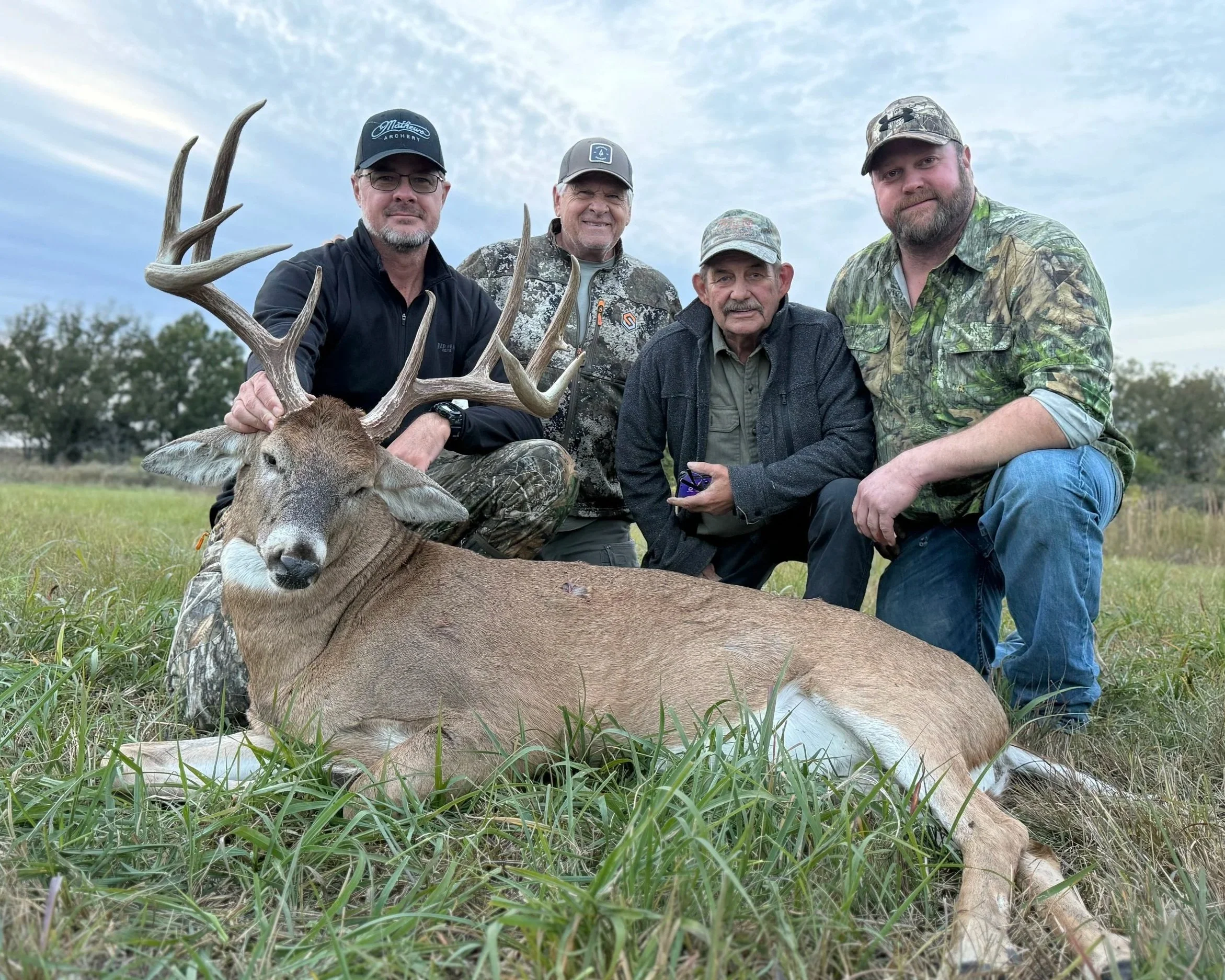 Four men posing with a slain deer in an open field under a cloudy sky.