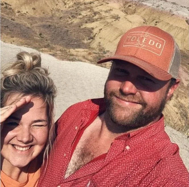 A smiling woman with blonde hair and a man with a beard wearing a red shirt and orange cap outdoors in a desert landscape.