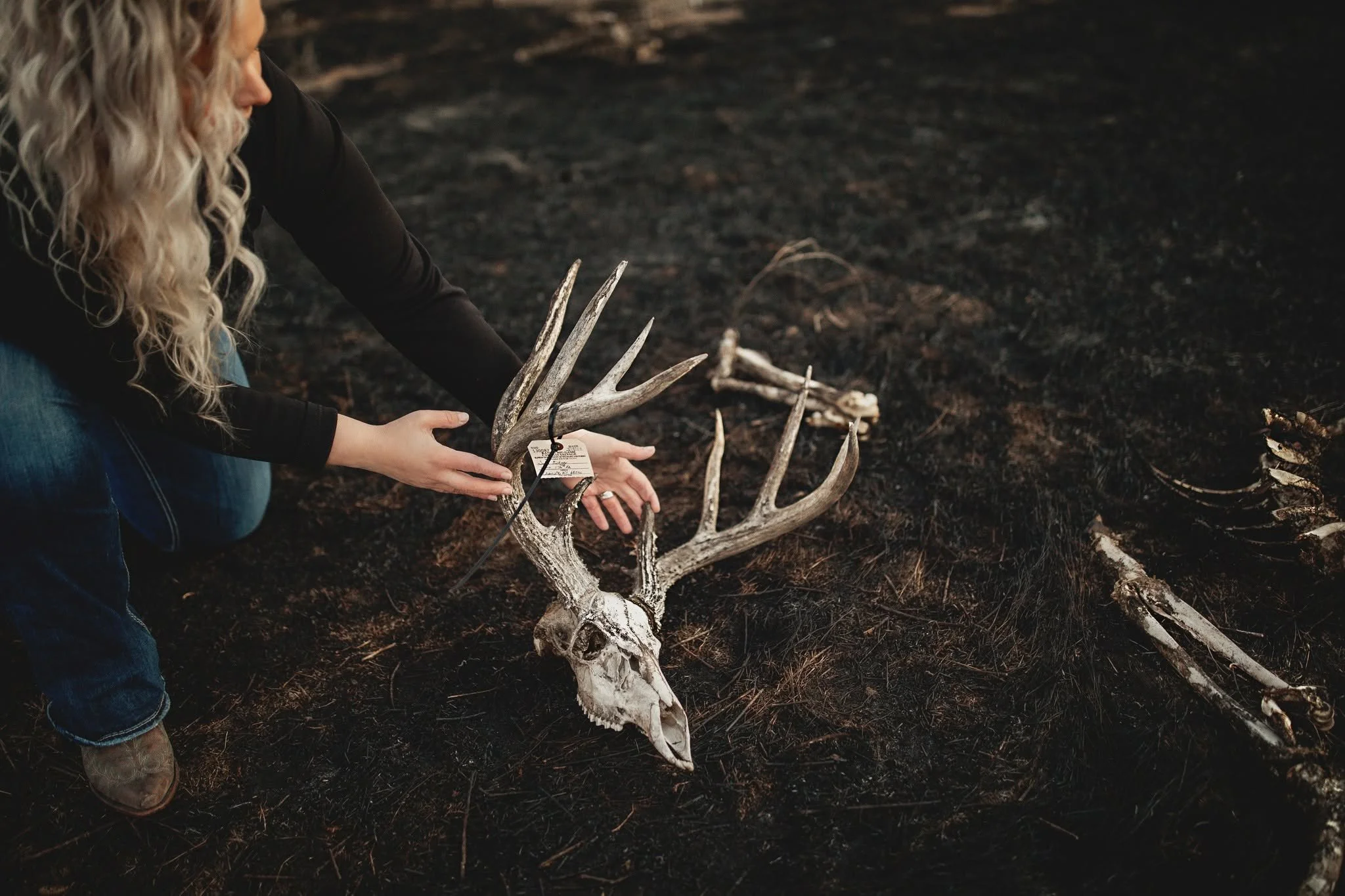 A person with long curly blonde hair wearing blue jeans and cowboy boots kneels on dark, ashy ground, arranging a white animal skull with antlers, possibly a deer, on the ground.