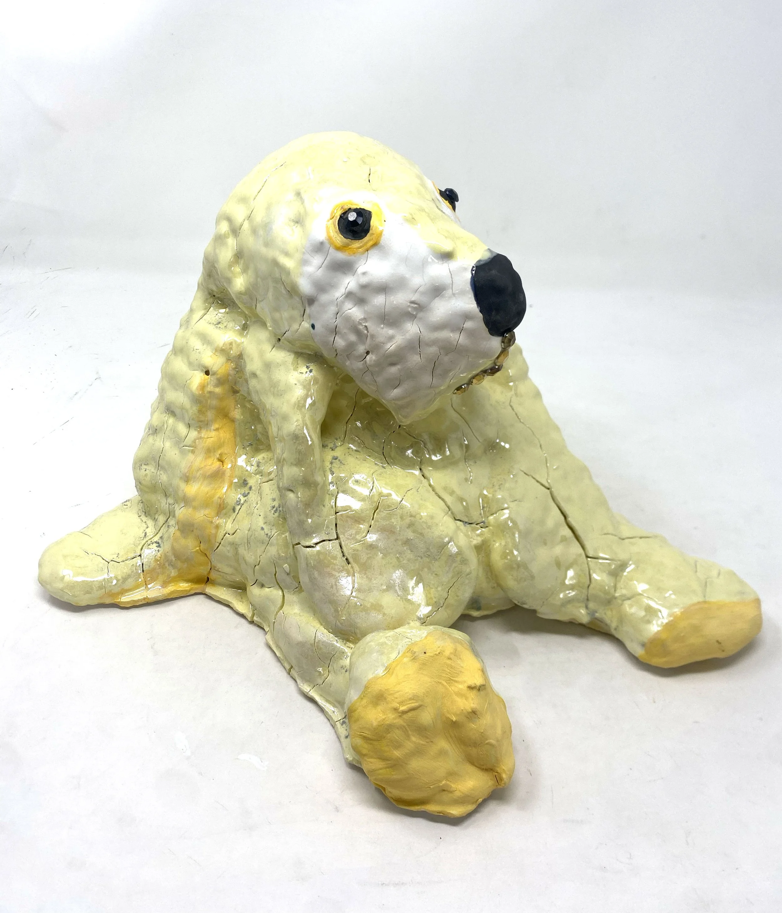 A sculpted figure resembling a bunny with a yellow body and a white face, made of a material like papier-mâché or painted ceramic, with visible cracks and painted black eyes and nose, sitting on a white surface against a plain white background.