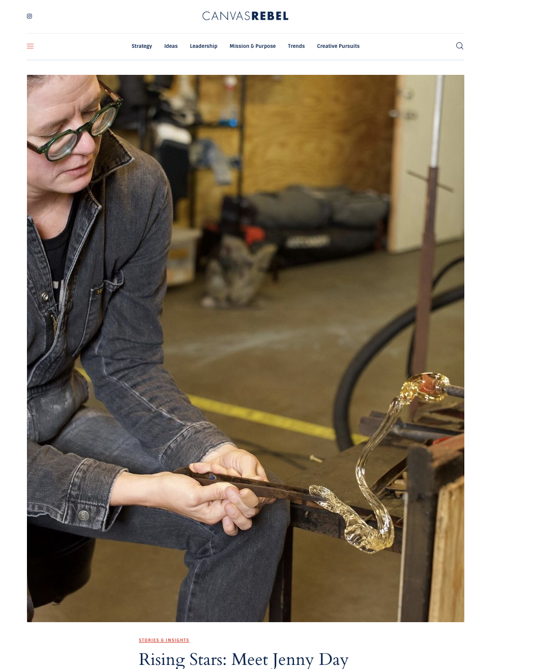 A person wearing glasses and a denim jacket working with glassblowing, shaping molten glass into a curved form in a workshop.
