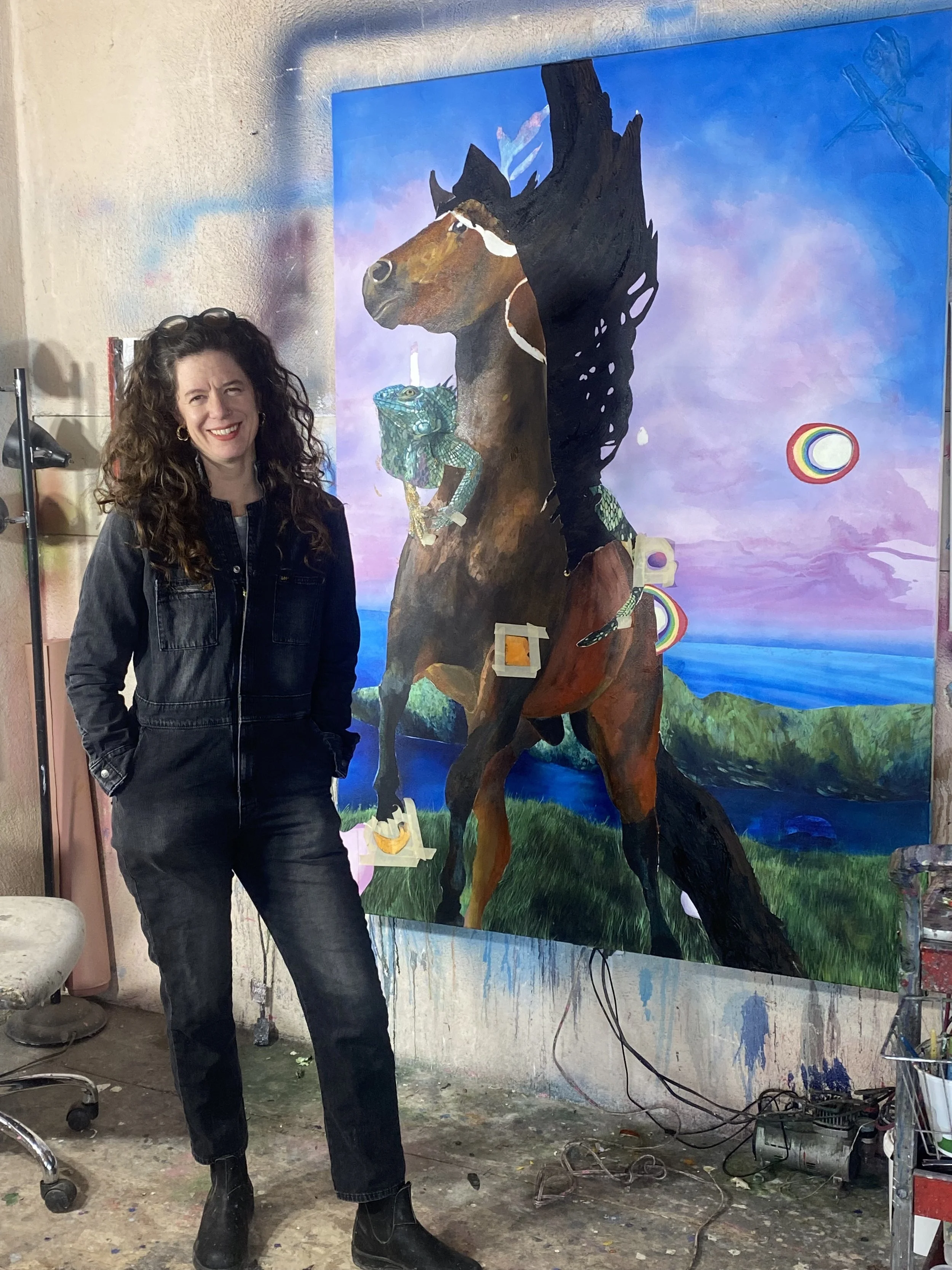A woman with curly hair, wearing glasses on her head, a denim jacket, and black pants, stands in front of a colorful painting of horses and dragons, in an art studio with paint splatters and tools visible.