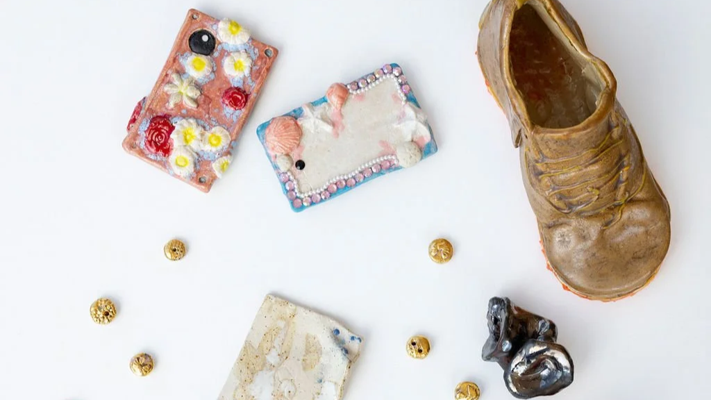 A variety of decorative and small objects on a white surface, including two decorated ceramic or clay plaques with floral and animal designs, a single brown and black shoe, a metallic figurine, and several small gold beads.
