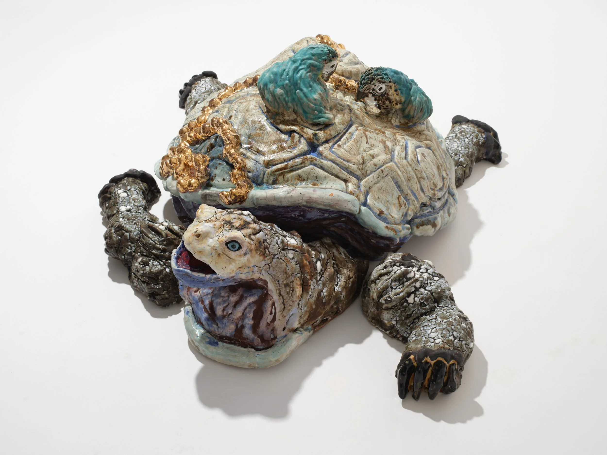 A sculpture of a turtle made with various textured and colorful ceramics and metals, featuring detailed skin patterns and a gold chain across its back, set against a plain white background.