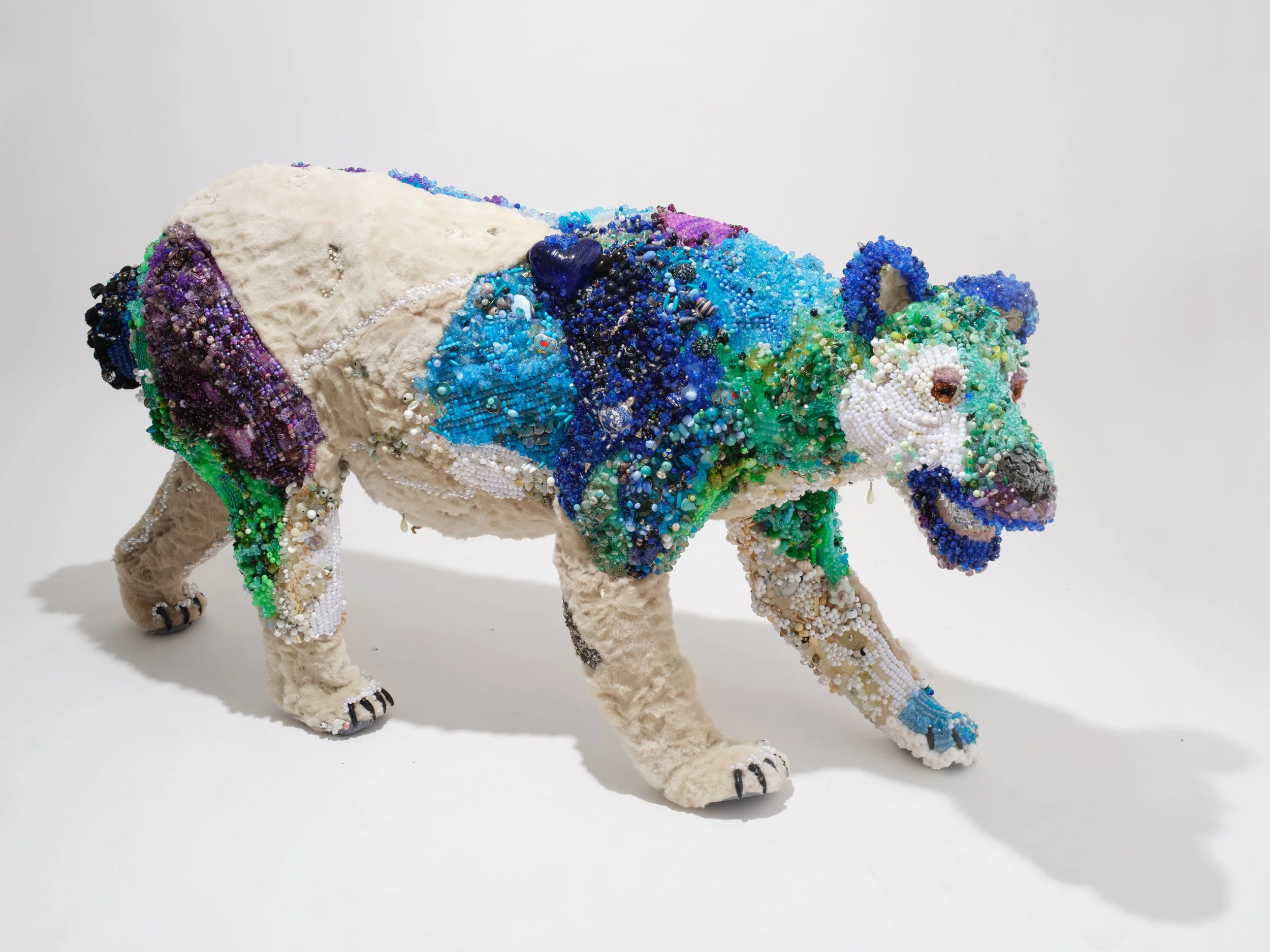 A sculpture of a bear, covered in colorful beads and fabric, with a white fluffy texture on its body and detailed facial features, set against a plain white background.