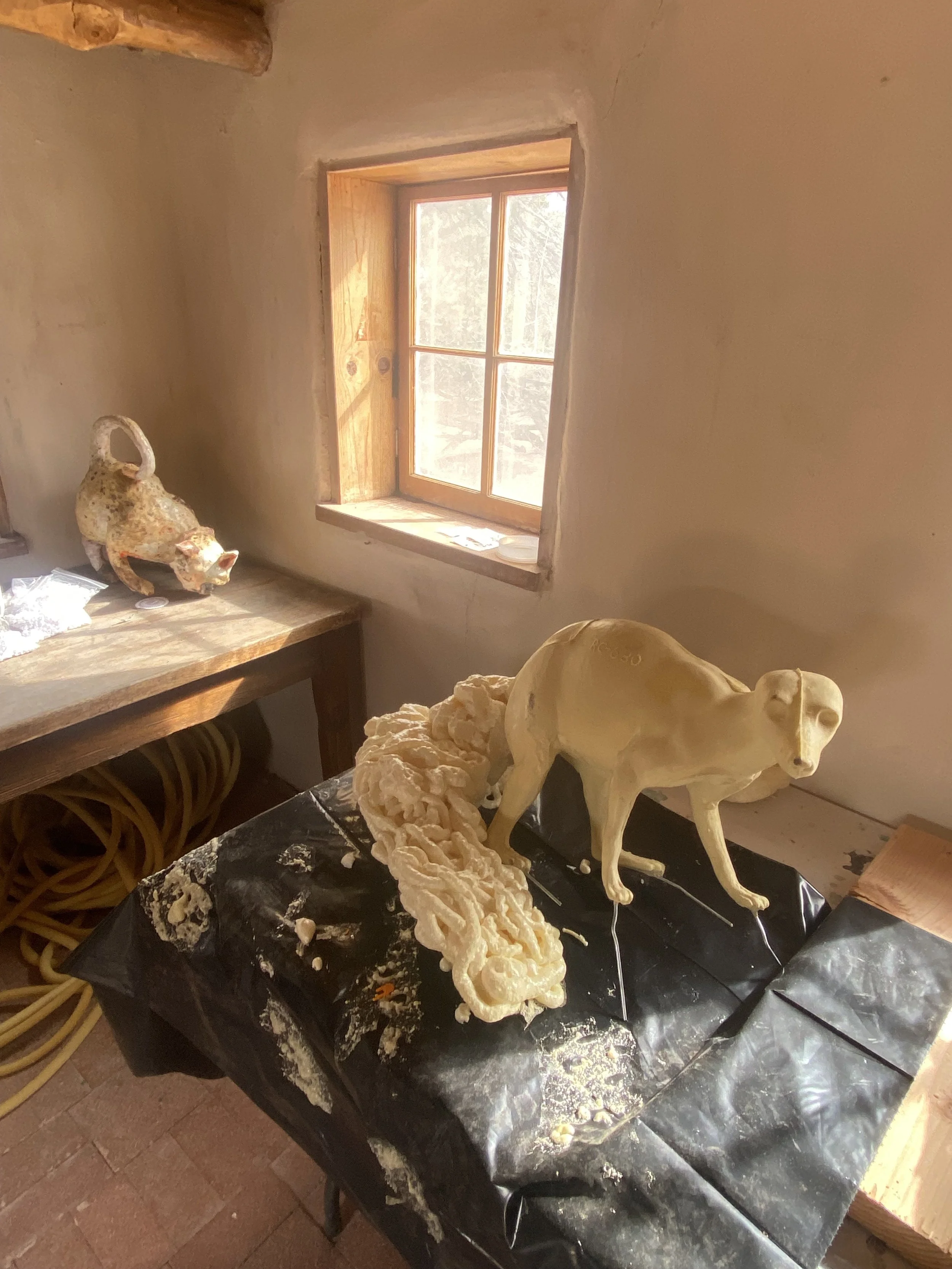An art studio workspace with sculptures and crafting materials on the table, illuminated by a window with wooden framing.