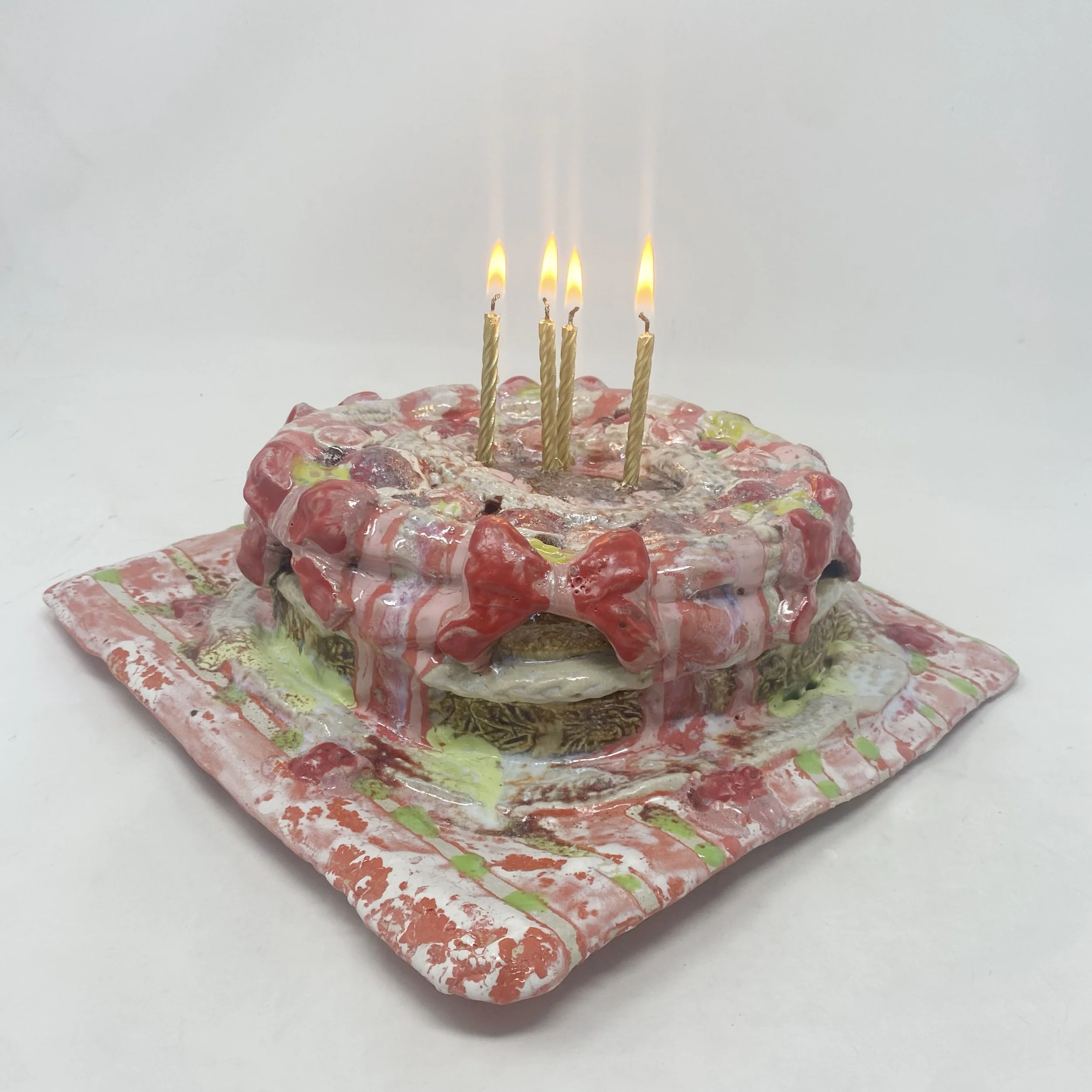 A colorful birthday cake with four lit candles on top, decorated with red, yellow, green, and white icing, on a multicolored ceramic plate against a plain white background.