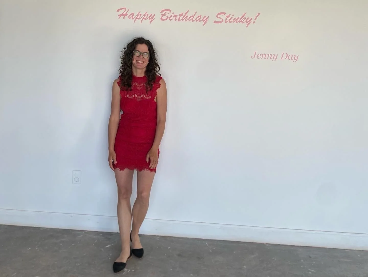 Woman in a red lace dress standing in front of a white wall with pink text that reads "Happy Birthday Stinky!" and "Jenny Day."