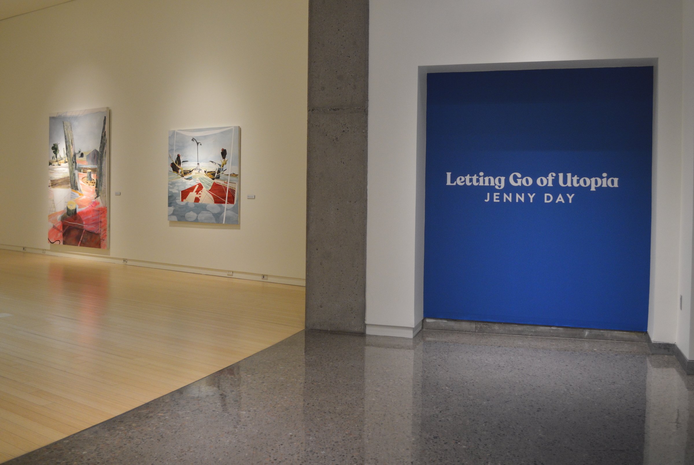 Museum wall with paintings and a blue sign reading 'Letting Go of Utopia' by Jenny Day.