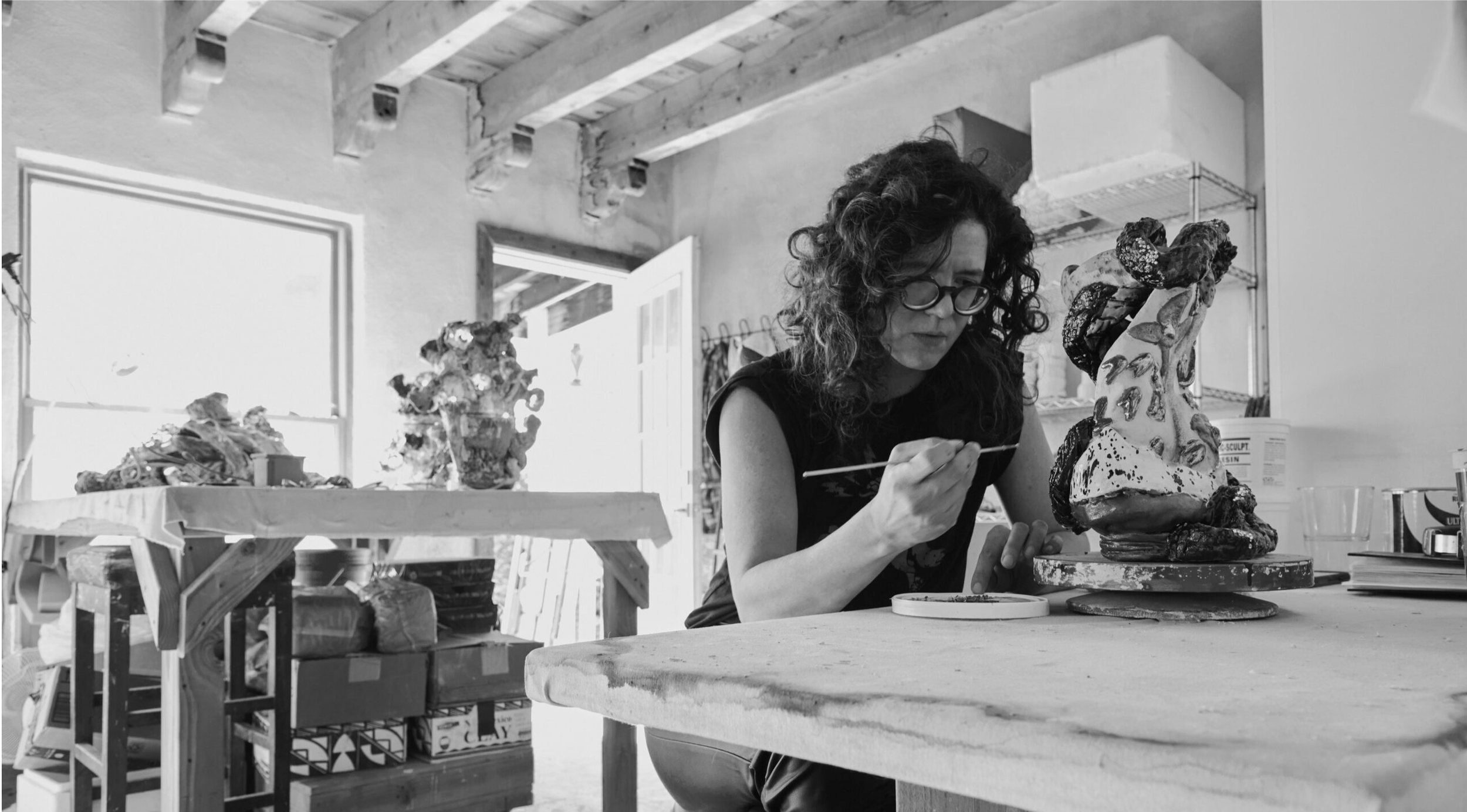 A woman with curly hair and glasses carefully paints a large abstract sculpture in an art studio.