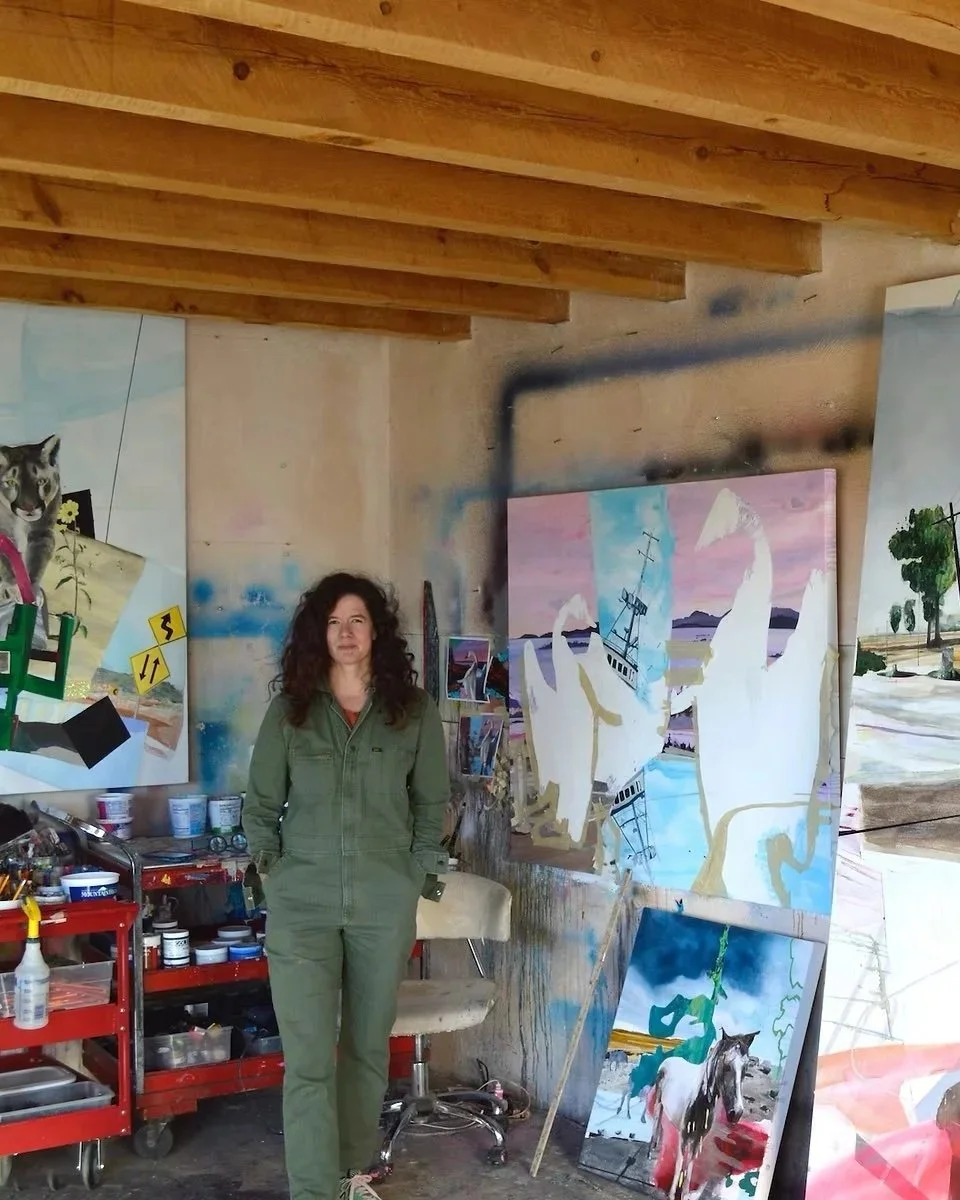 A woman in green overalls standing in an art studio with colorful paintings, including a large painting of a ship and white swans, and a red cart filled with art supplies.