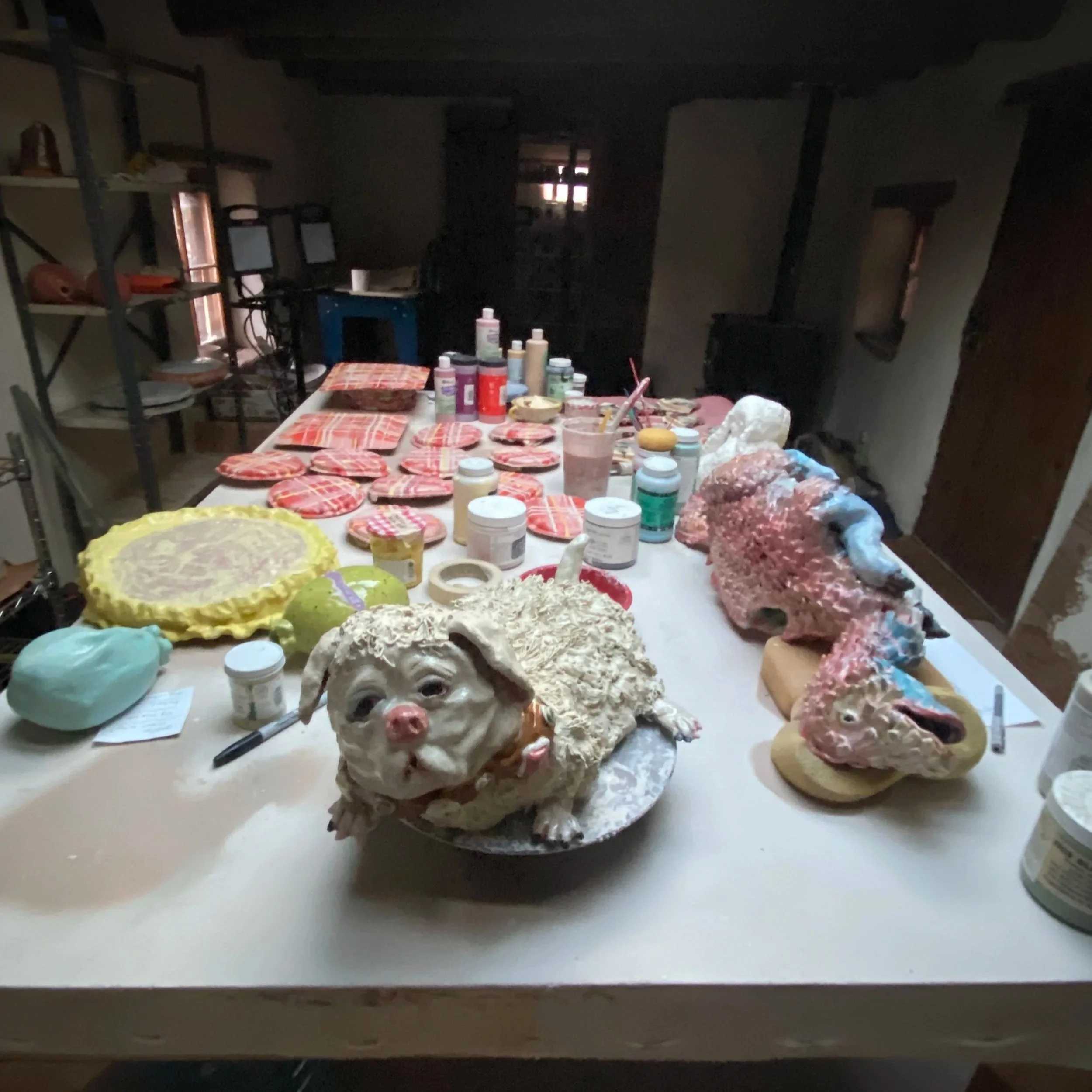 A dimly lit room with a table covered in various art supplies and crafts, including jars of paint, brushes, and decorated items. There are shelves with more items and a small window letting in some light.