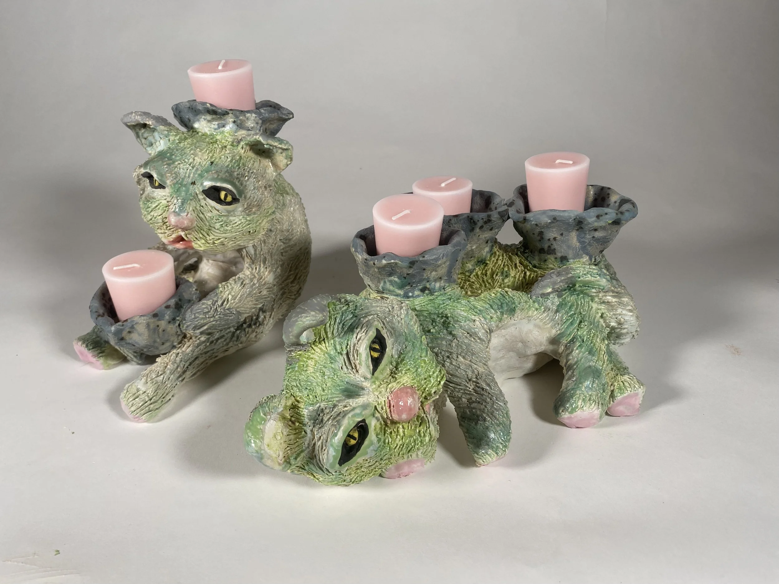Decorative ceramic candle holder shaped like a cat with pink candles on its back, green and yellow accents, set against a plain background.