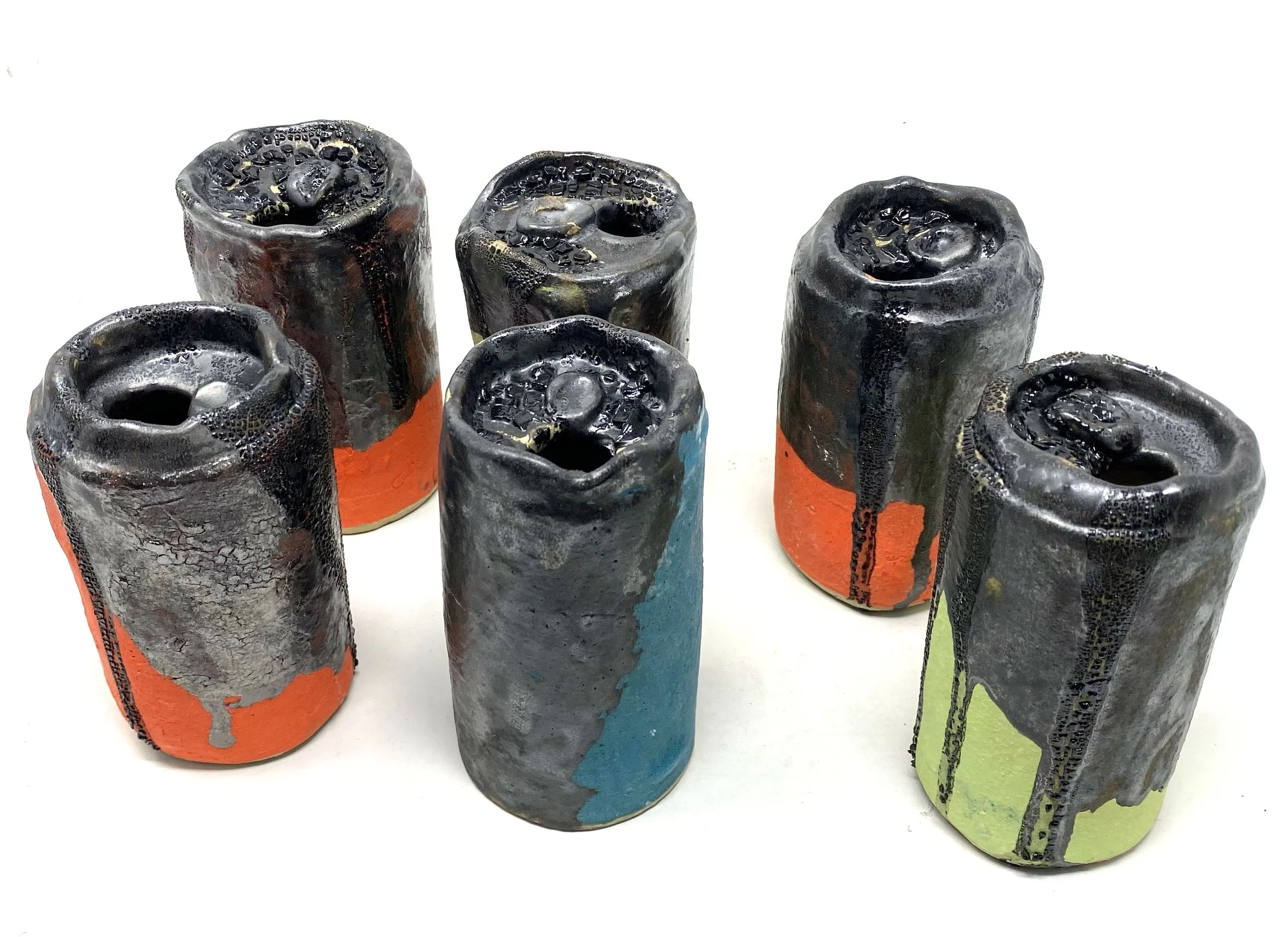 Six cylindrical objects with a textured, bubbly, black surface top, partially covered with colored paint in orange, blue, green, and red.