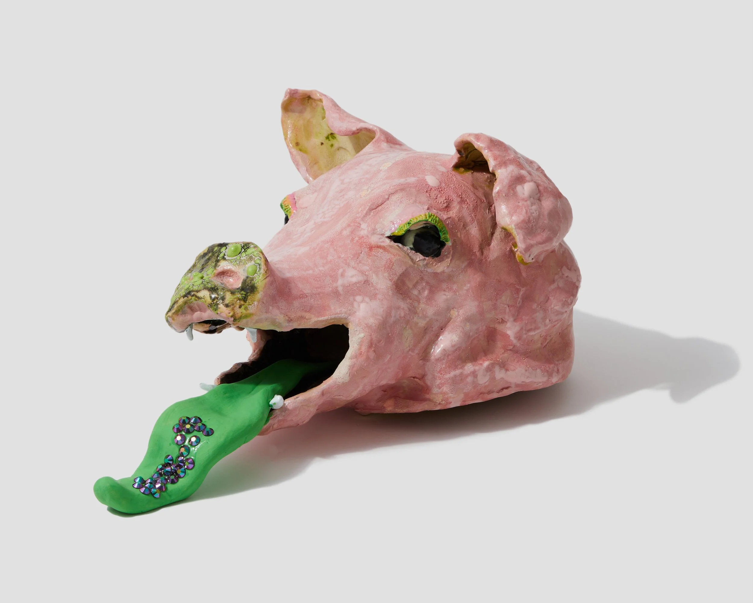 A sculpture of a pink pig's head with a green tongue, decorated with small shiny beads, on a white background.