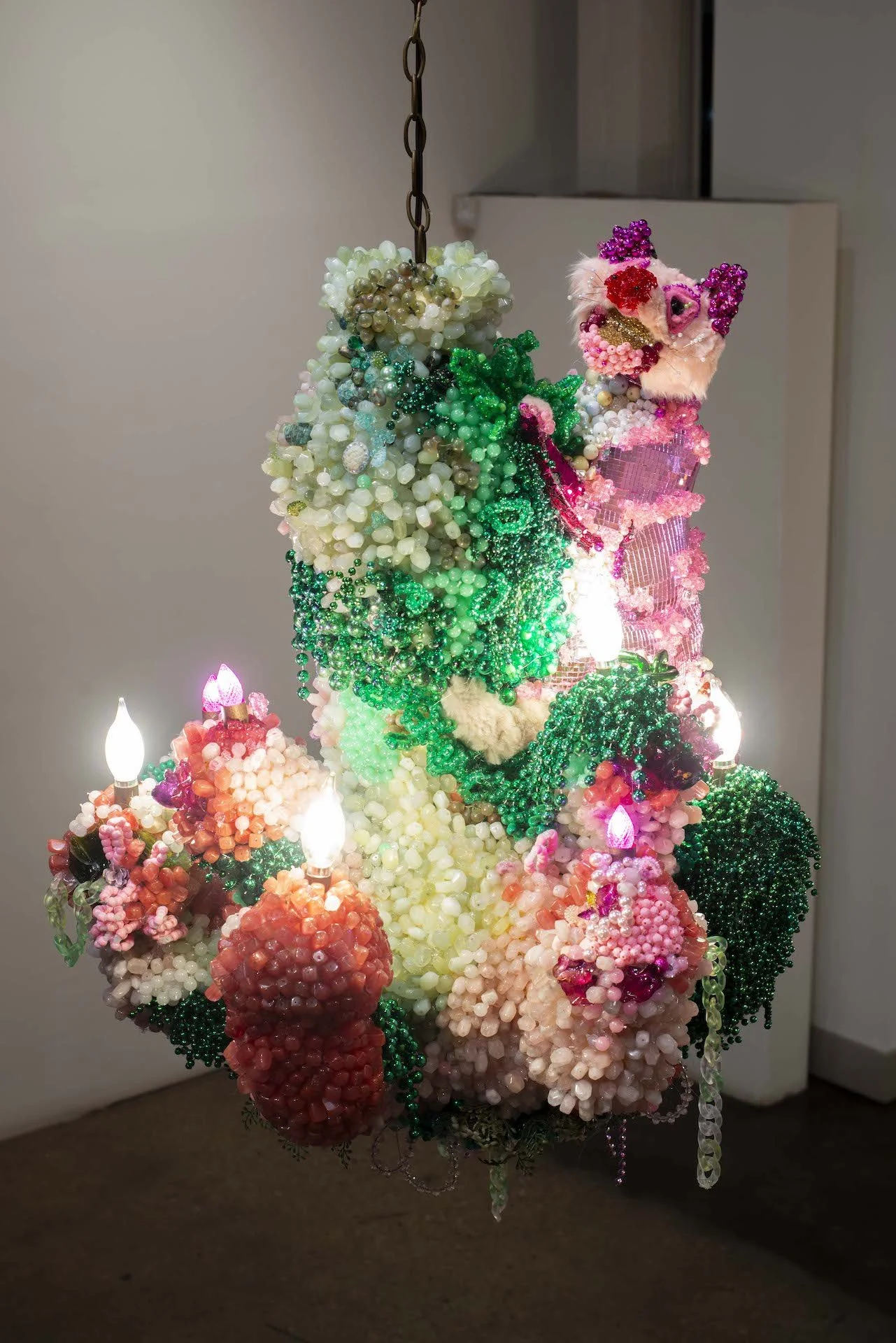 Colorful, intricate art installation resembling a sculpture made of various beads, fabrics, and small lights, with a mix of pink, green, white, and red elements, displayed indoors against a neutral background.