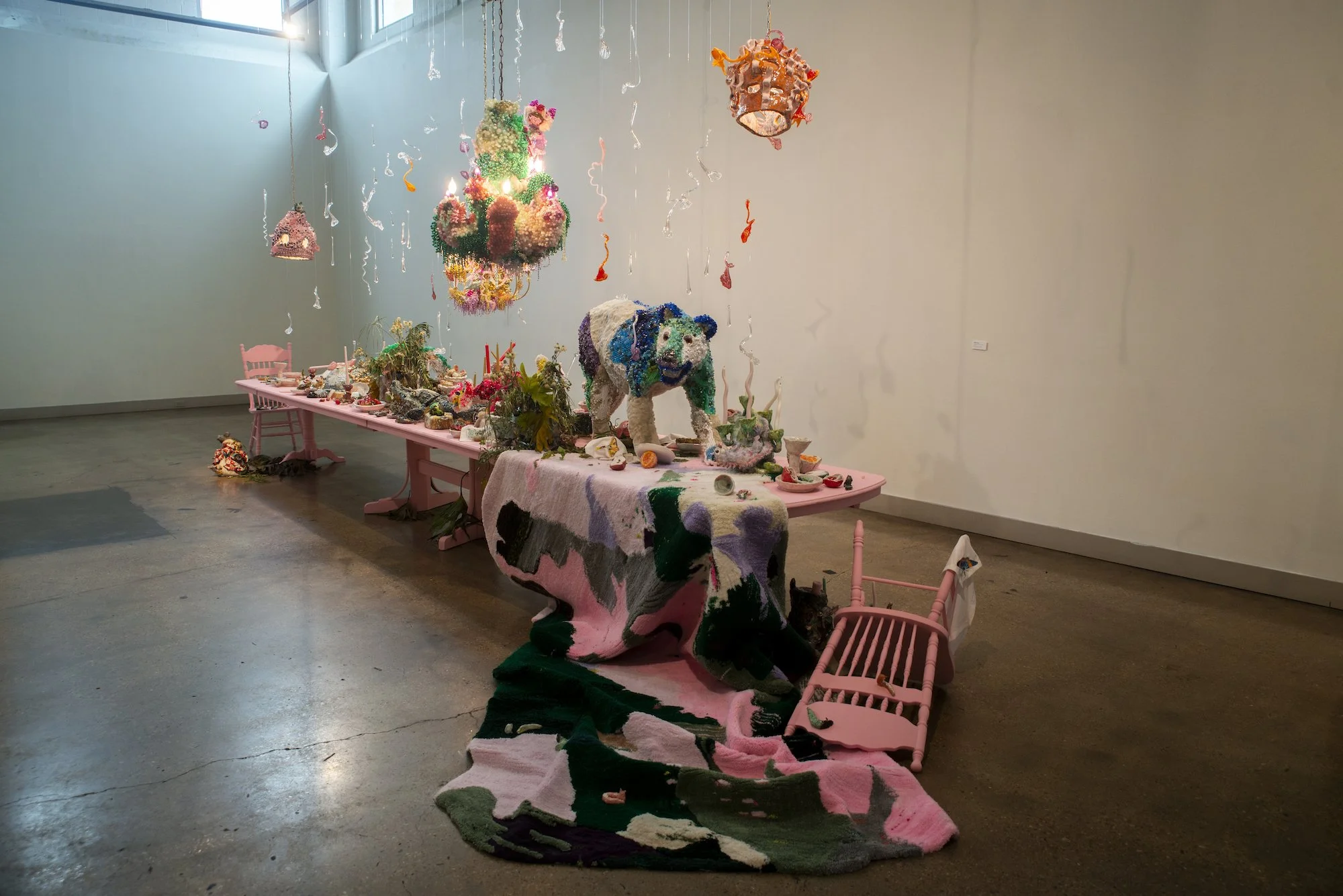 A colorful art installation featuring a large panda sculpture made of various textured materials, positioned on a pink table covered with a multicolored cloth. Hanging above are several suspended, whimsical objects, and the scene is set in a gallery with plain walls and a concrete floor.