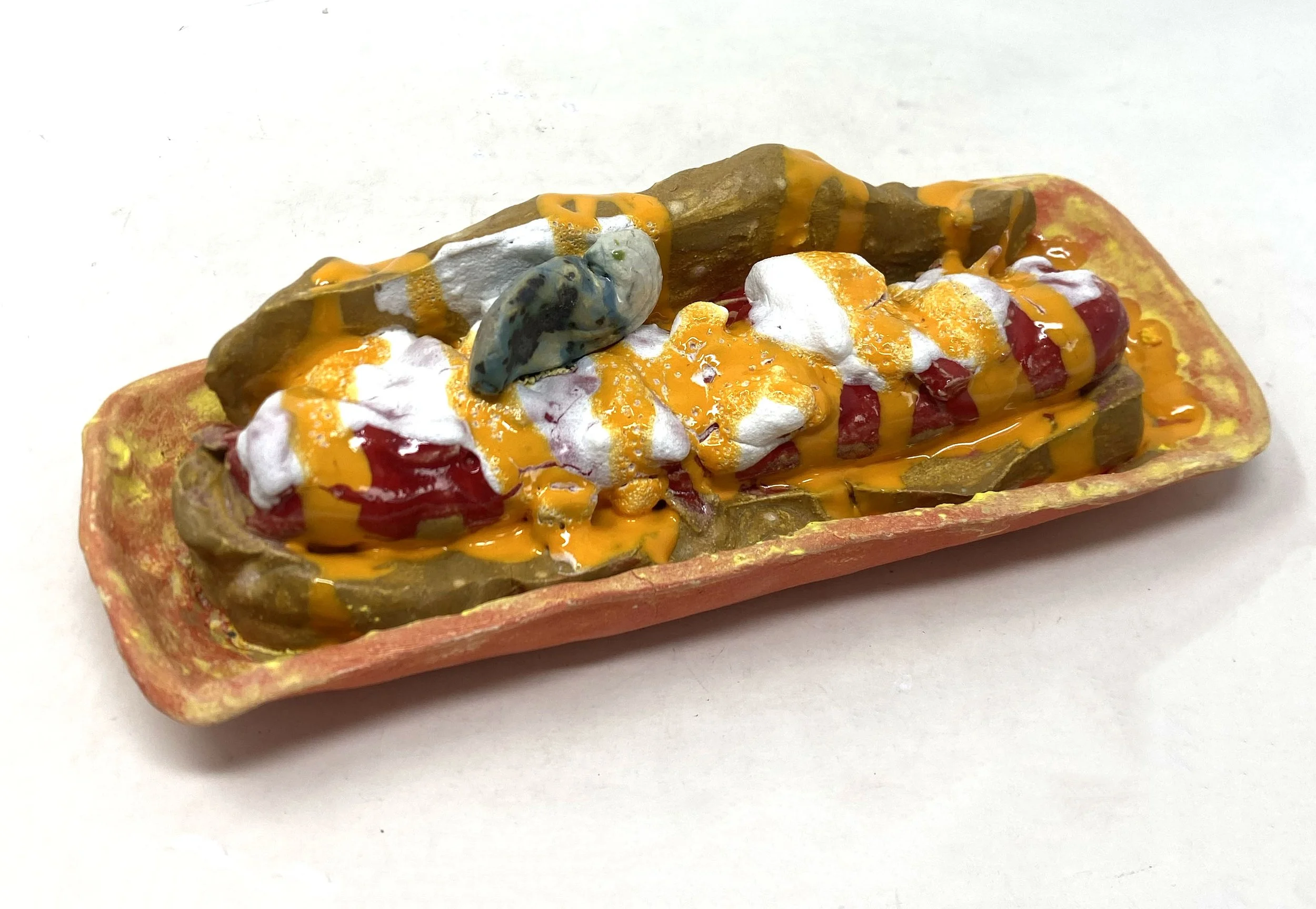 Close-up of a decorative item or sculpture resembling a hotdog with toppings, placed in a rectangular dish.