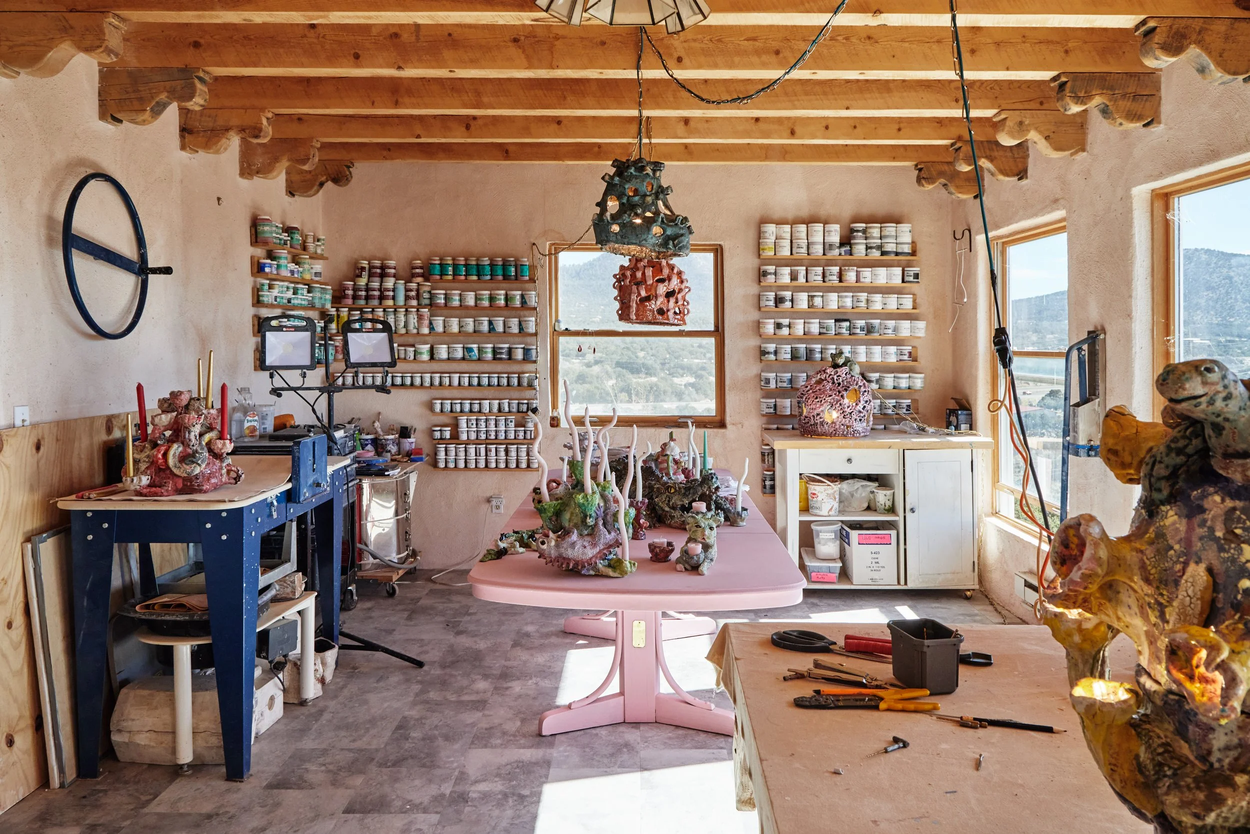 Art studio with shelves of paint jars on beige walls, wooden ceiling beams, a pink table with sculptures, and windows showing a scenic view outside.