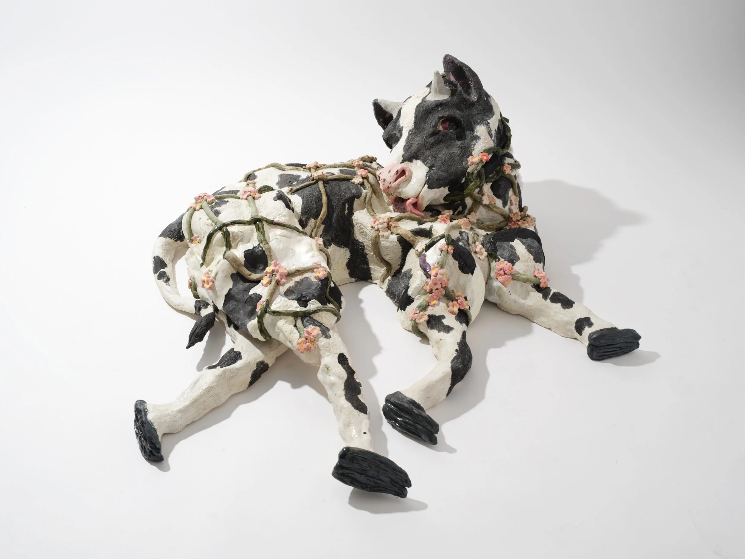 A ceramic sculpture of a cow lying down, with black and white patches, pink flowers on its body, and black hooves and eyes, set against a plain white background.