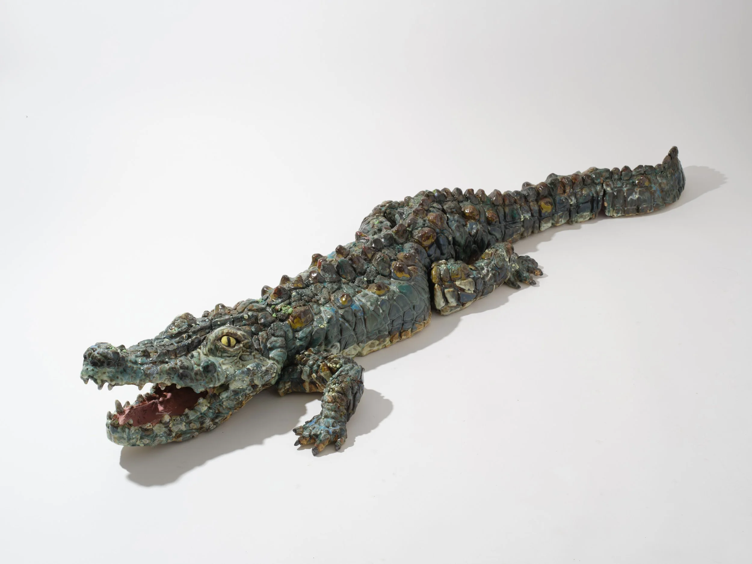A realistic sculpture of a crocodile made from colorful, textured materials, positioned on a plain white background with its mouth slightly open.