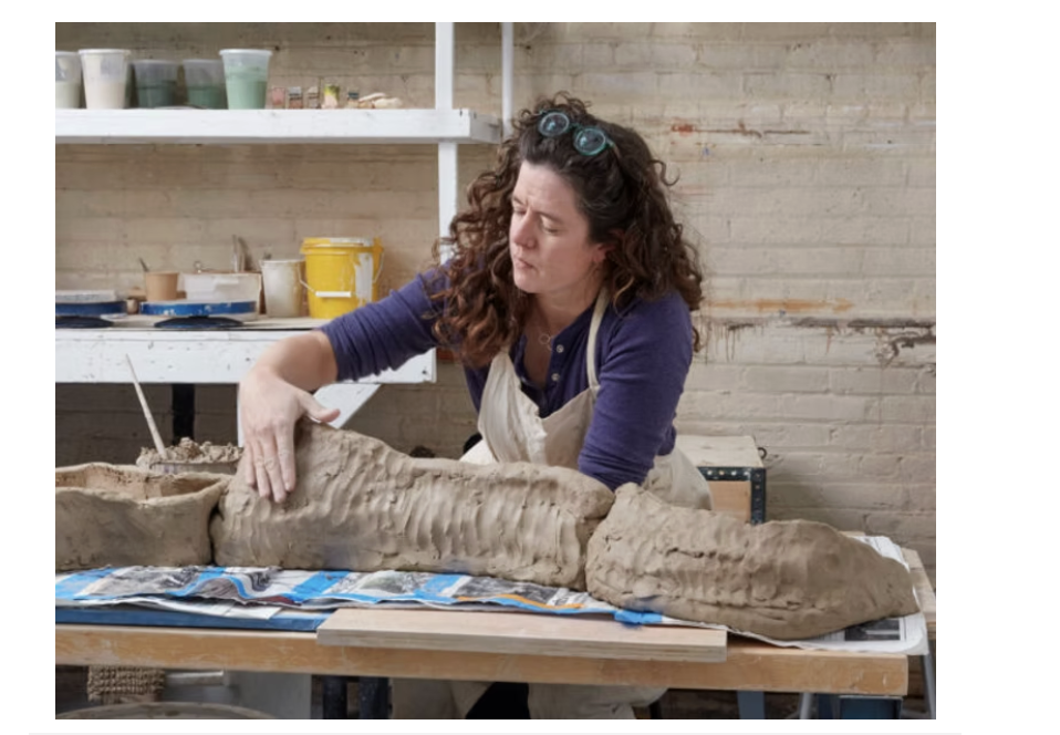 Woman working on a large clay sculpture in an art studio, with shelves of art supplies in the background.