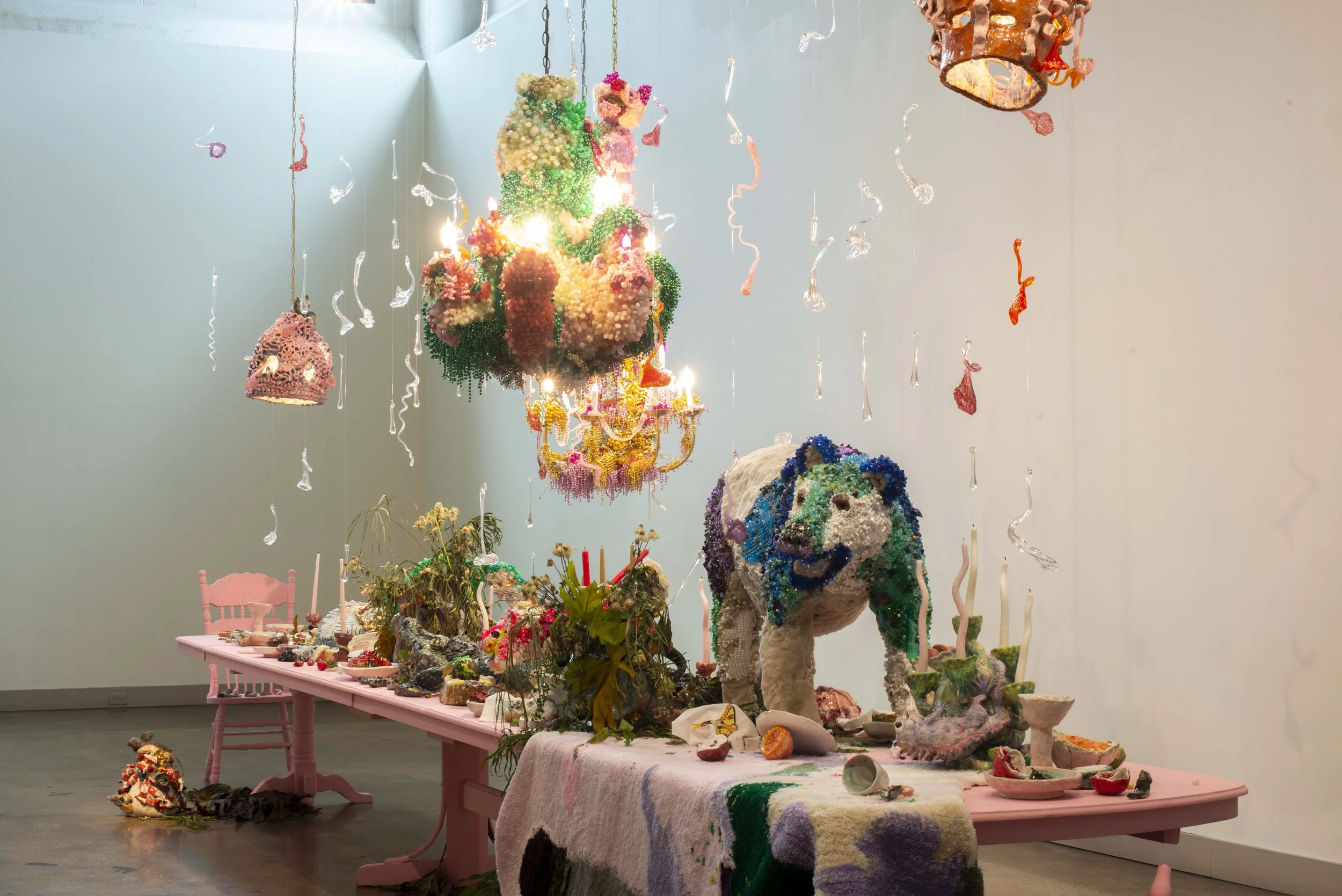 Art installation with colorful, sculptural objects resembling abstract figures and animals, suspended and arranged on a pink table and hanging from the ceiling, in an art gallery setting.