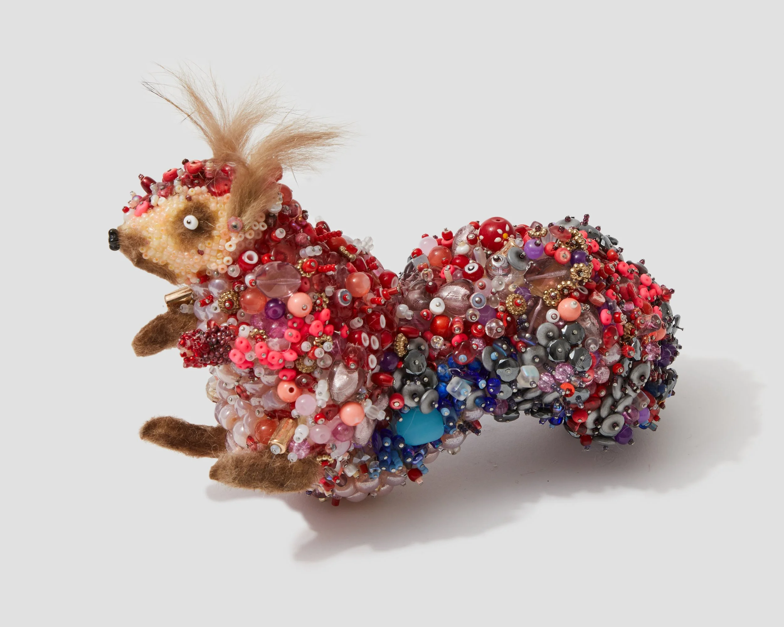 A sculpture of a squirrel covered in multicolored beads, sequins, and embellishments, with a light brown tuft of fur on its head, set against a plain white background.