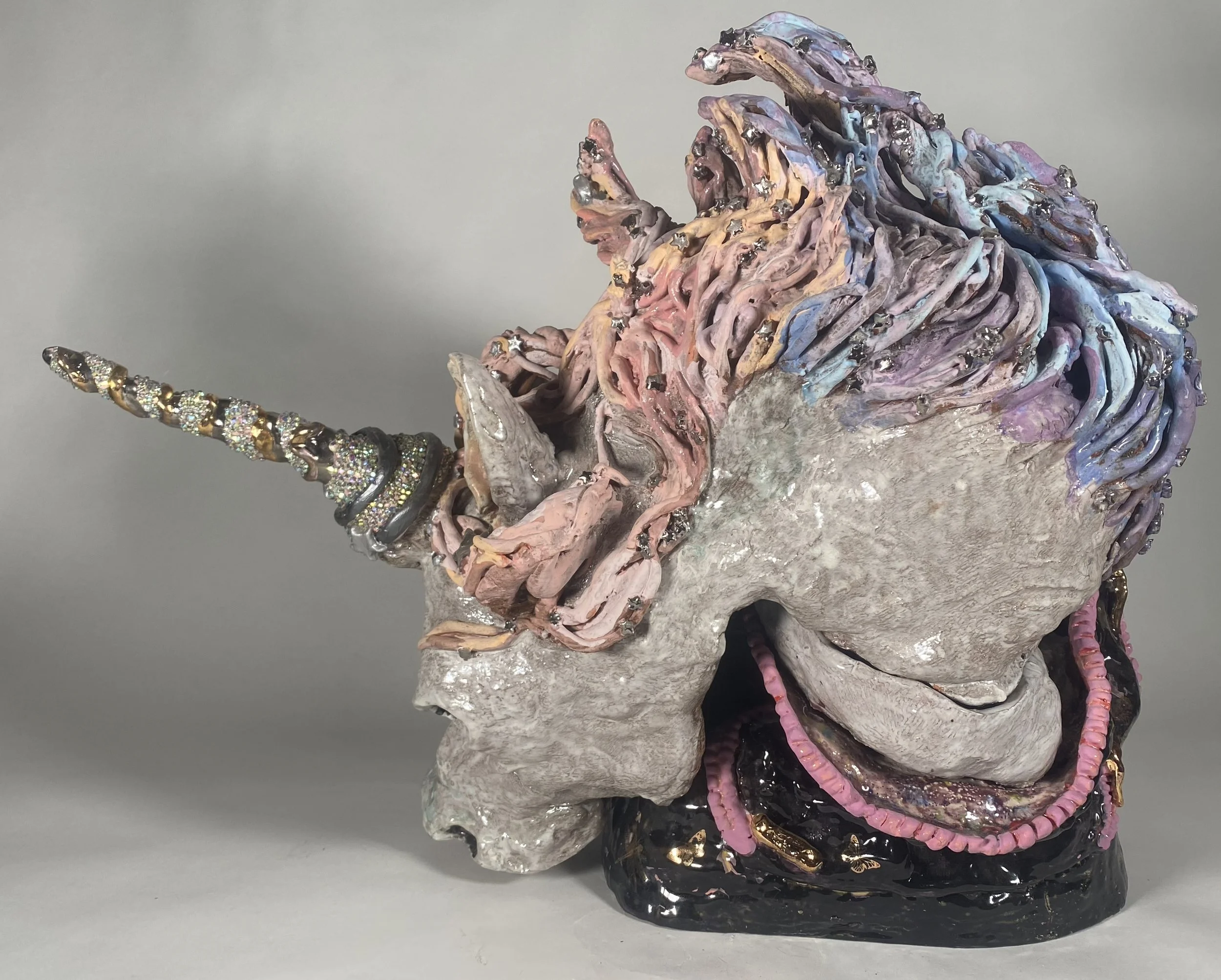 Sculpture of a unicorn head with a pastel color palette, decorated with rhinestones, set against a gray background.