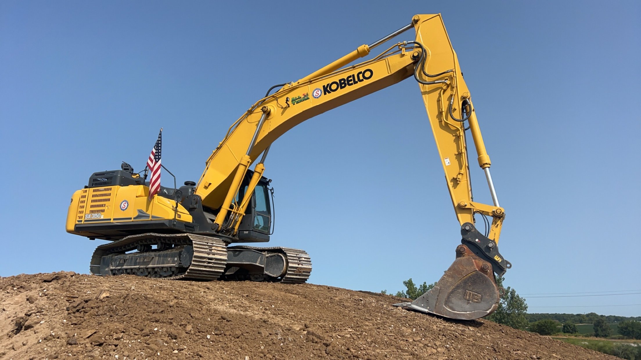 Yellow Kobelco excavator on a dirt mound with a blue sky background.