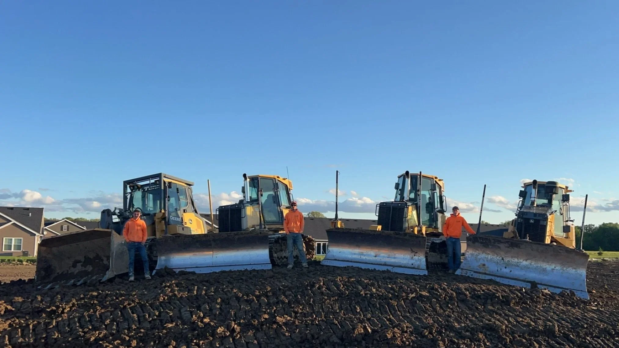 Three construction workers stand on a dirt ground in front of three large bulldozers, with a residential neighborhood and blue sky in the background.
